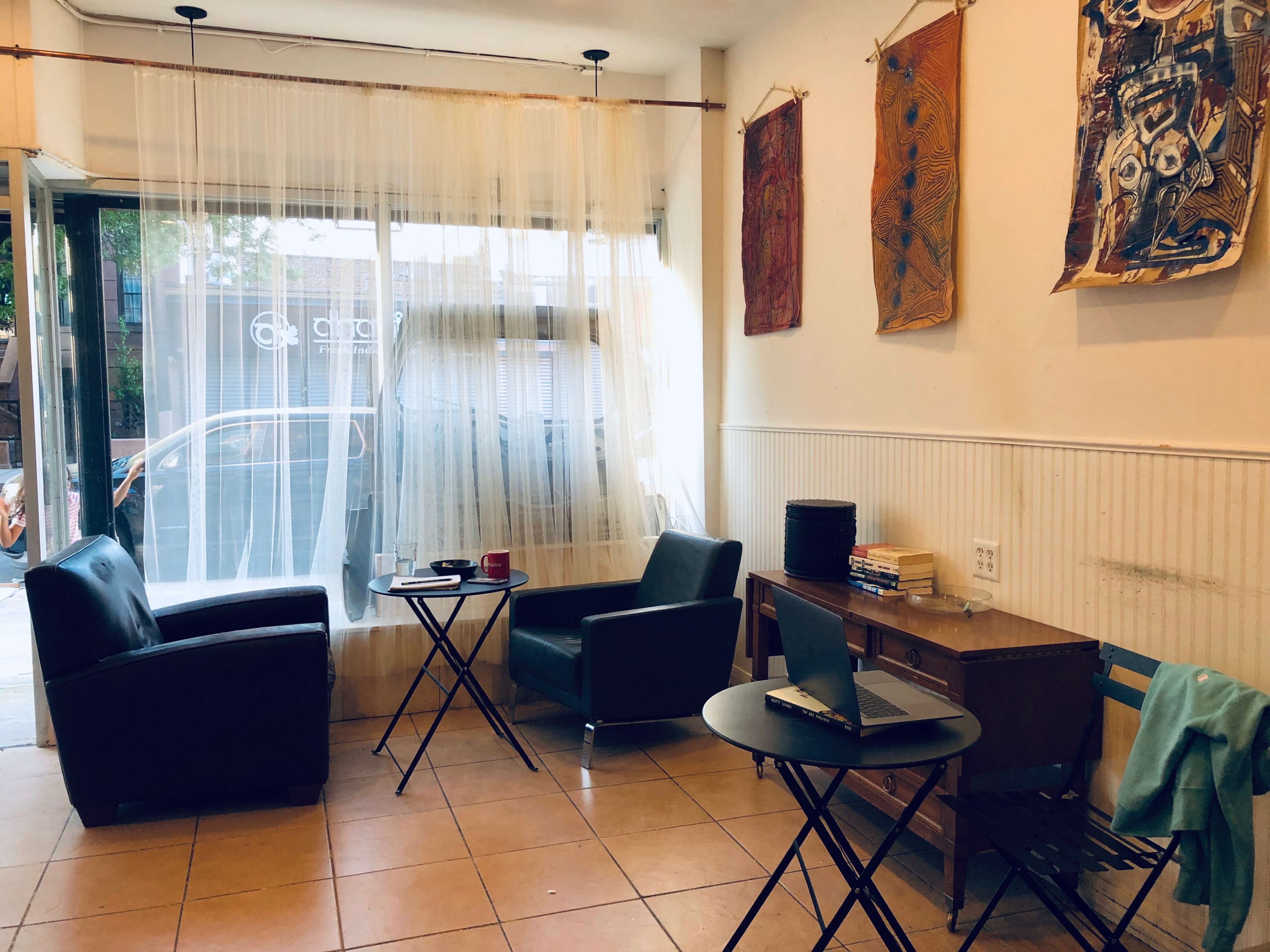 A small, minimalist café interior featuring two chairs, a small round table, a desk with a laptop, and decorative wall hangings.