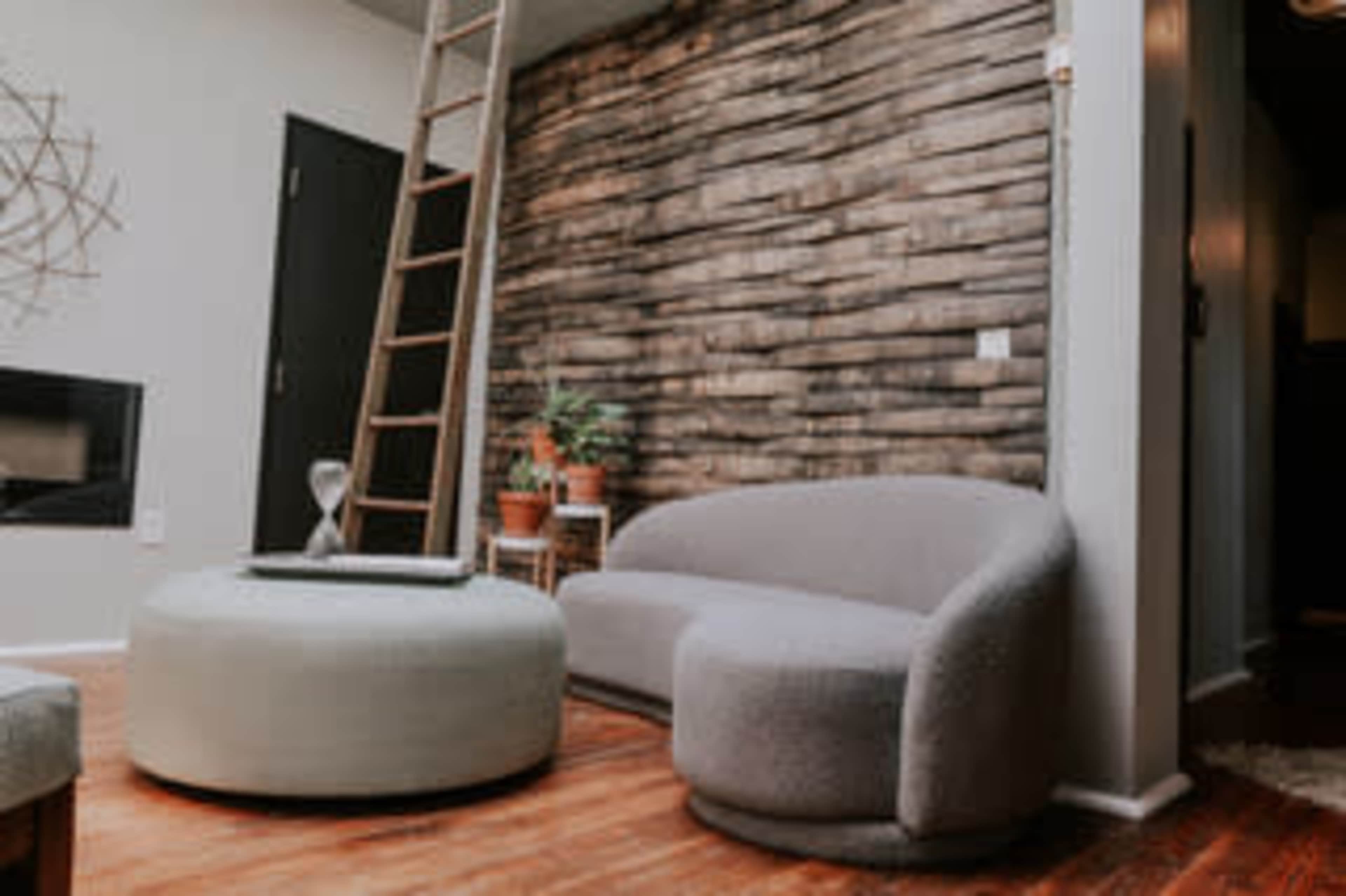 A modern living room with a curved gray sofa, a round ottoman, a wooden ladder against a textured wall, and potted plants.