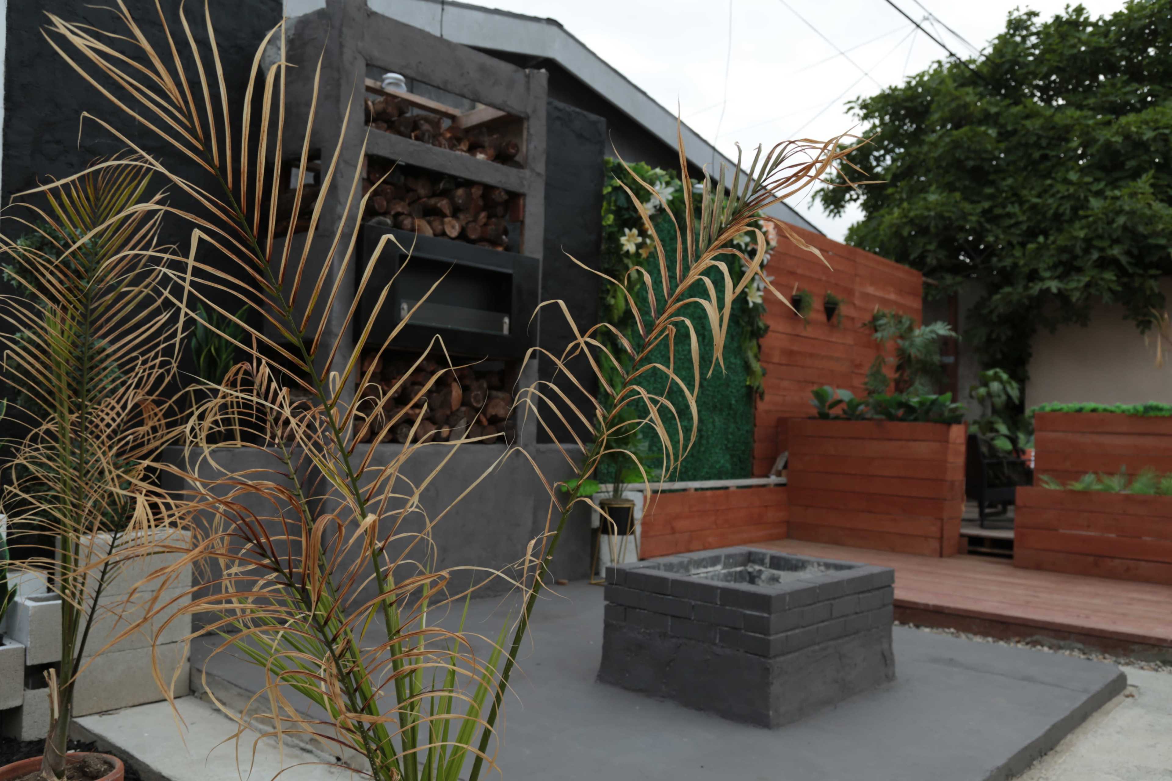 A modern outdoor patio area featuring a stone fire pit, vertical wood storage, and landscaped greenery.