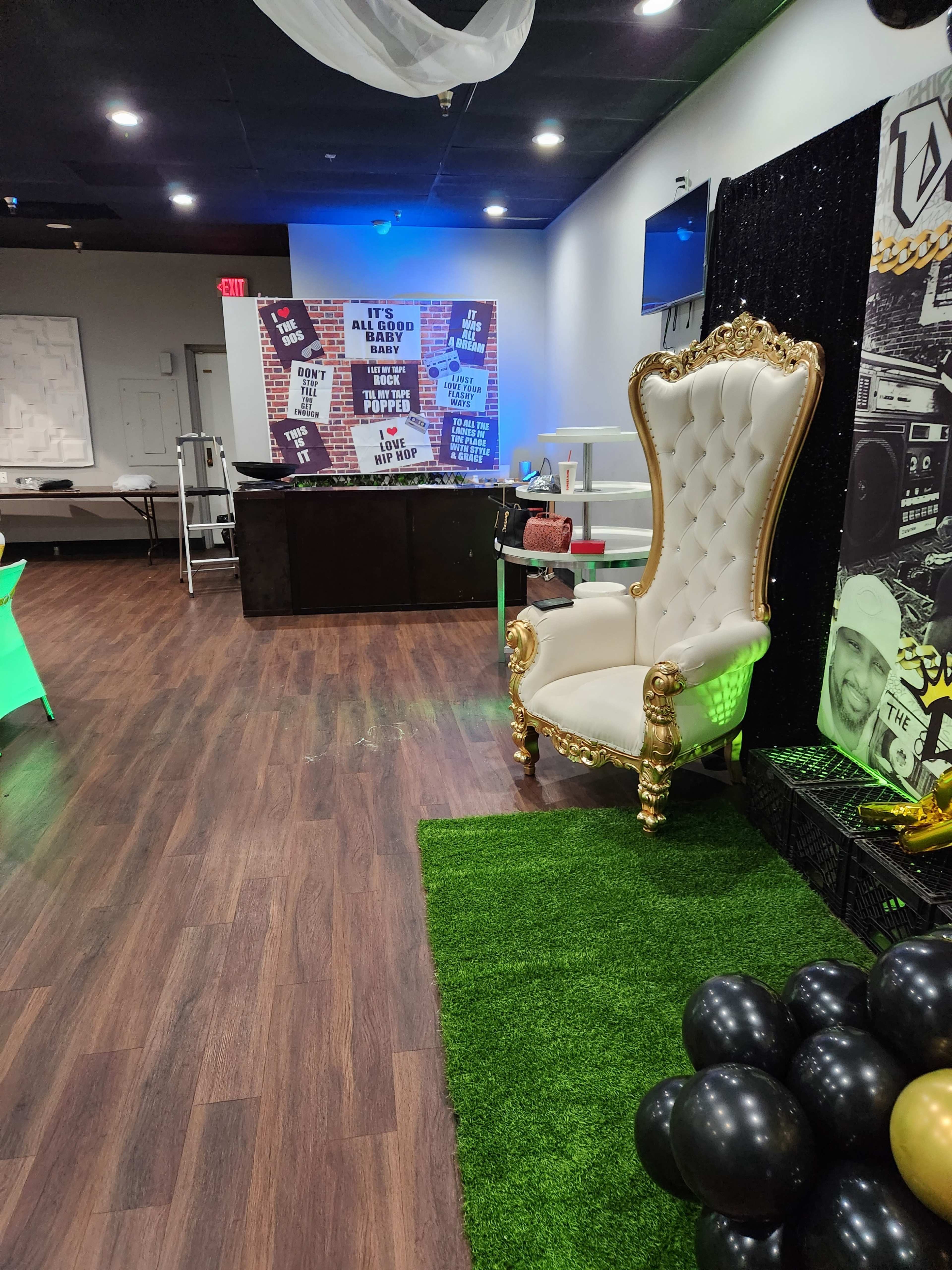The image shows a decorated event space featuring a golden throne chair, green grass-like flooring, and a backdrop with colorful graphics and text.