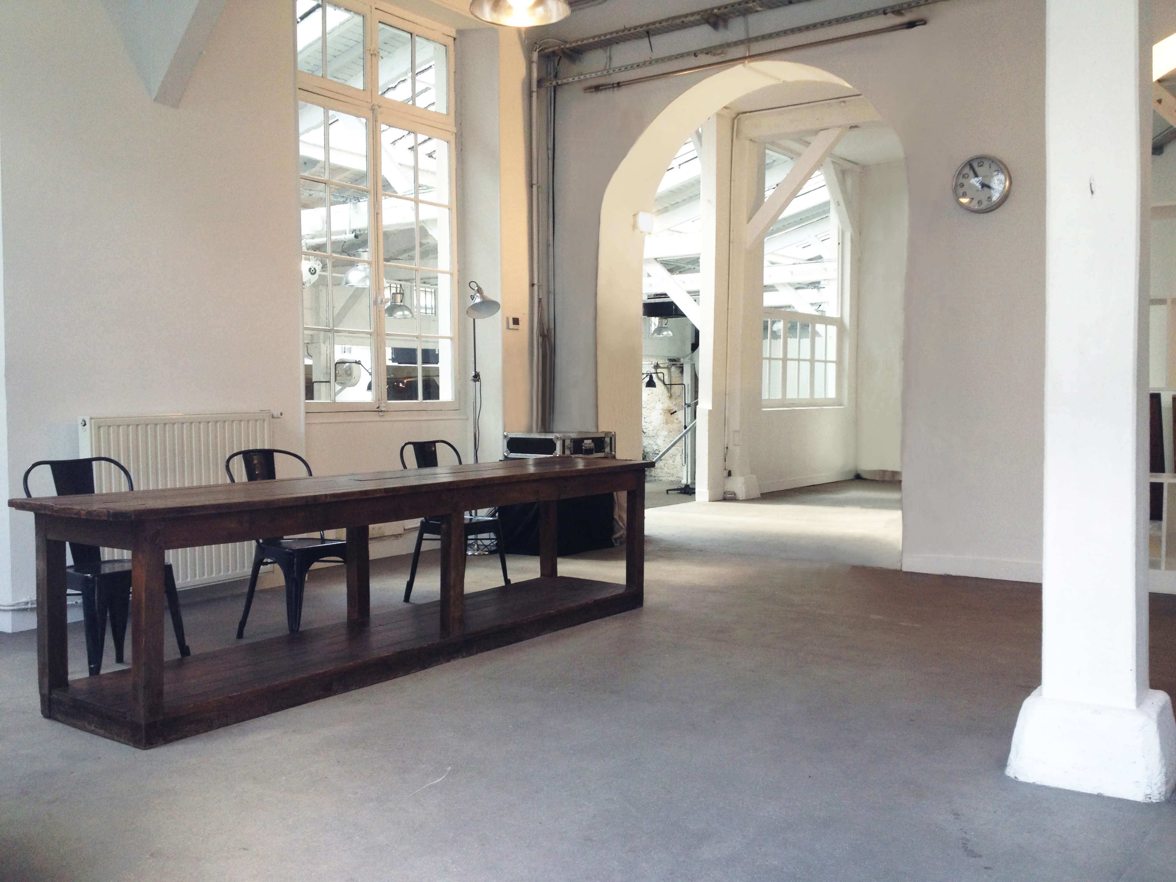 A spacious, bright room with a wooden table and black chairs, featuring large windows and an archway leading to another area.