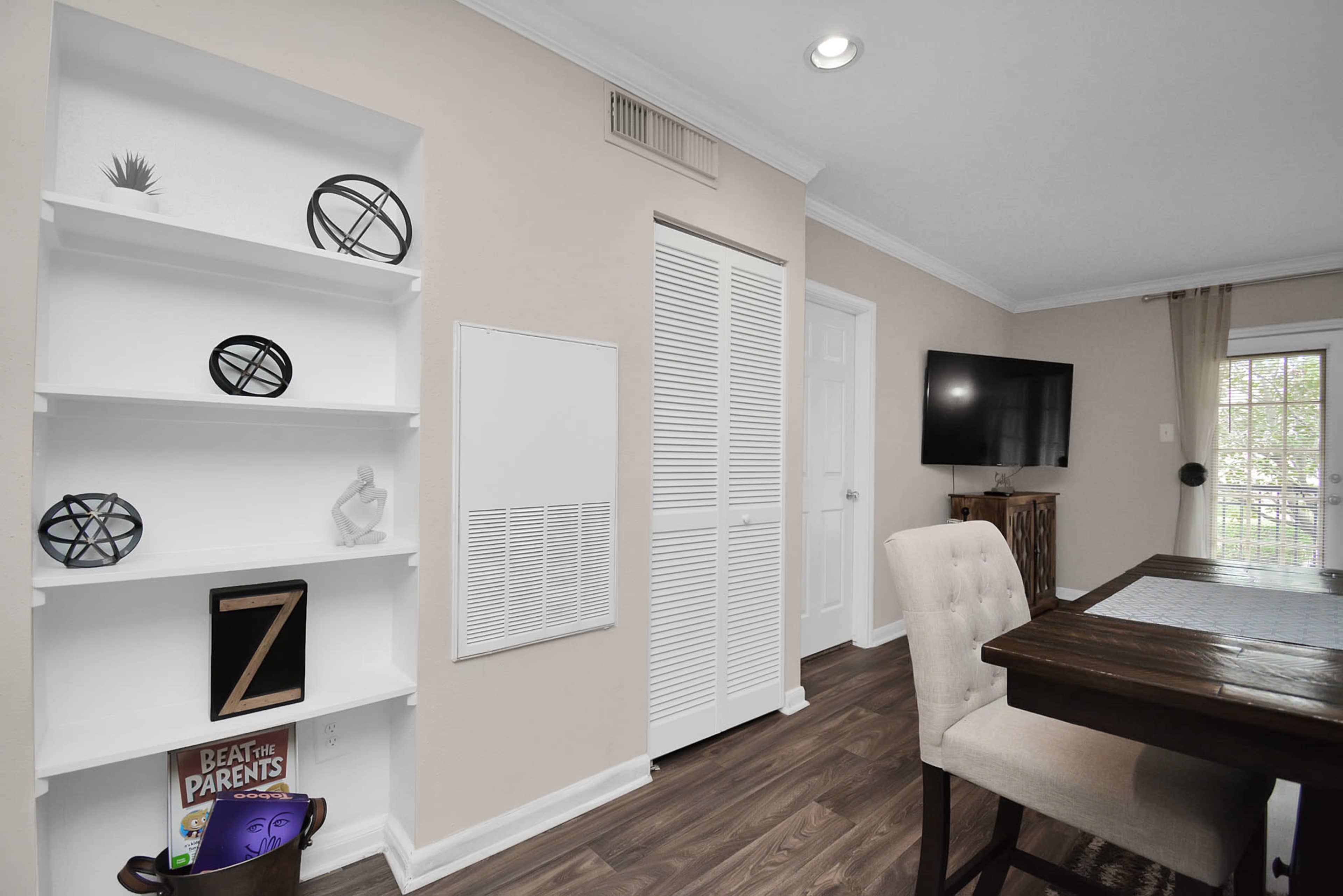 A modern interior featuring a shelf with decorative objects, a wooden dining table, and a television mounted on the wall, with light-colored walls and hardwood flooring.