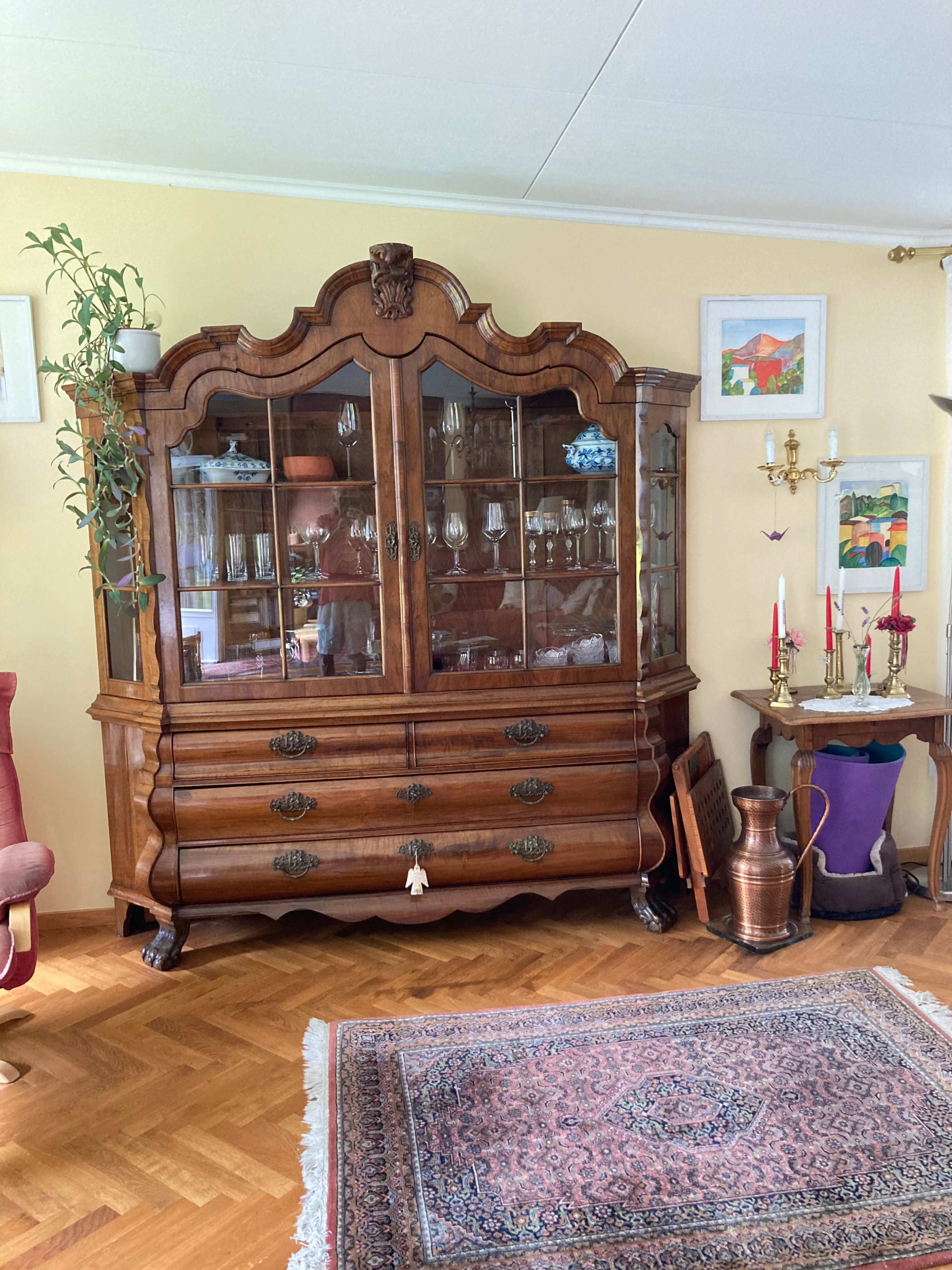 A wooden display cabinet with glass doors, a set of drawers below, and decorative items, positioned in a room with a patterned rug and various artwork on the walls.