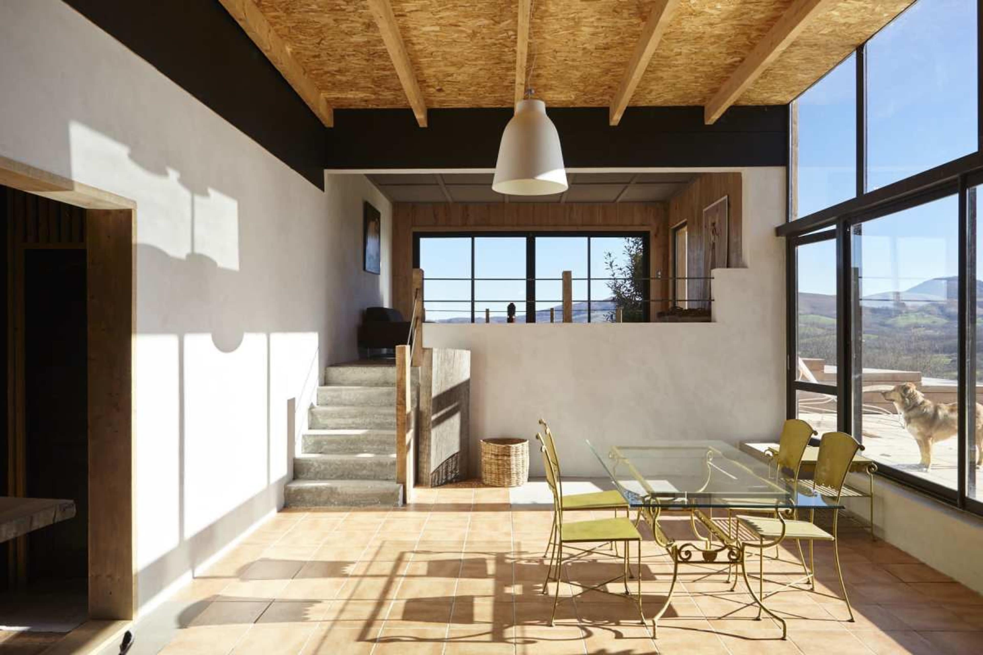 A modern interior features a glass dining table with yellow chairs, wooden accents, and large windows offering a view of the landscape.