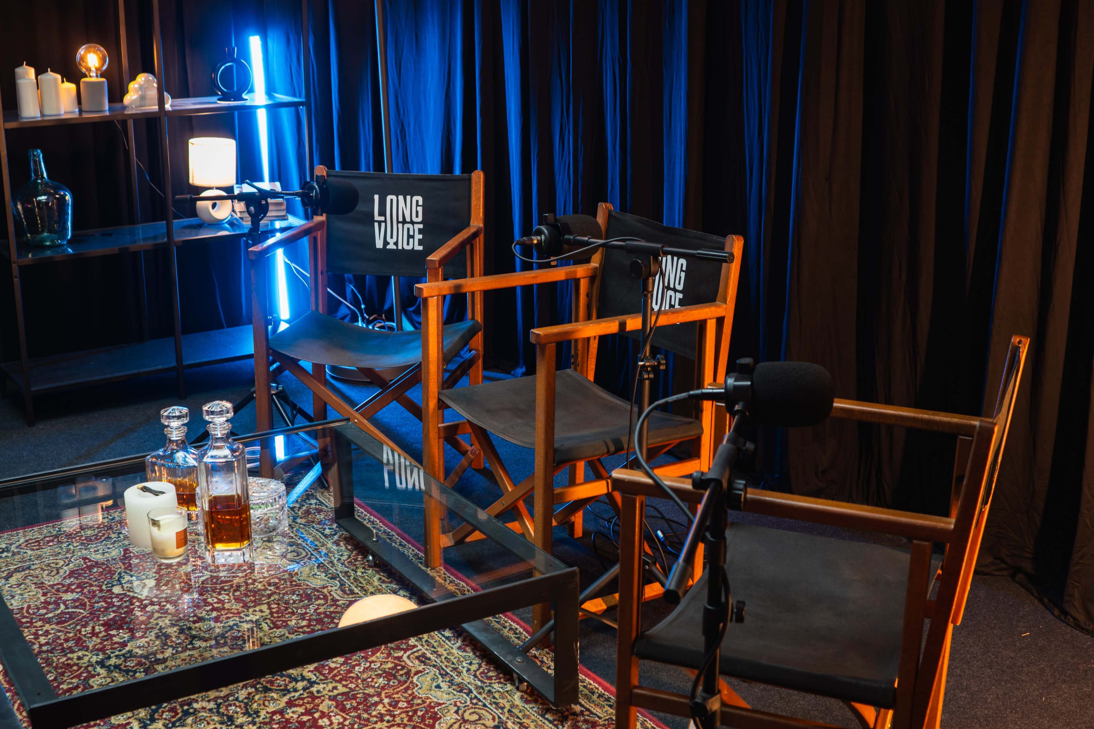 The image shows a studio setup with two director's chairs labeled "LONG" and "VOICE," microphones, a glass table with decorative items, and a dark backdrop.