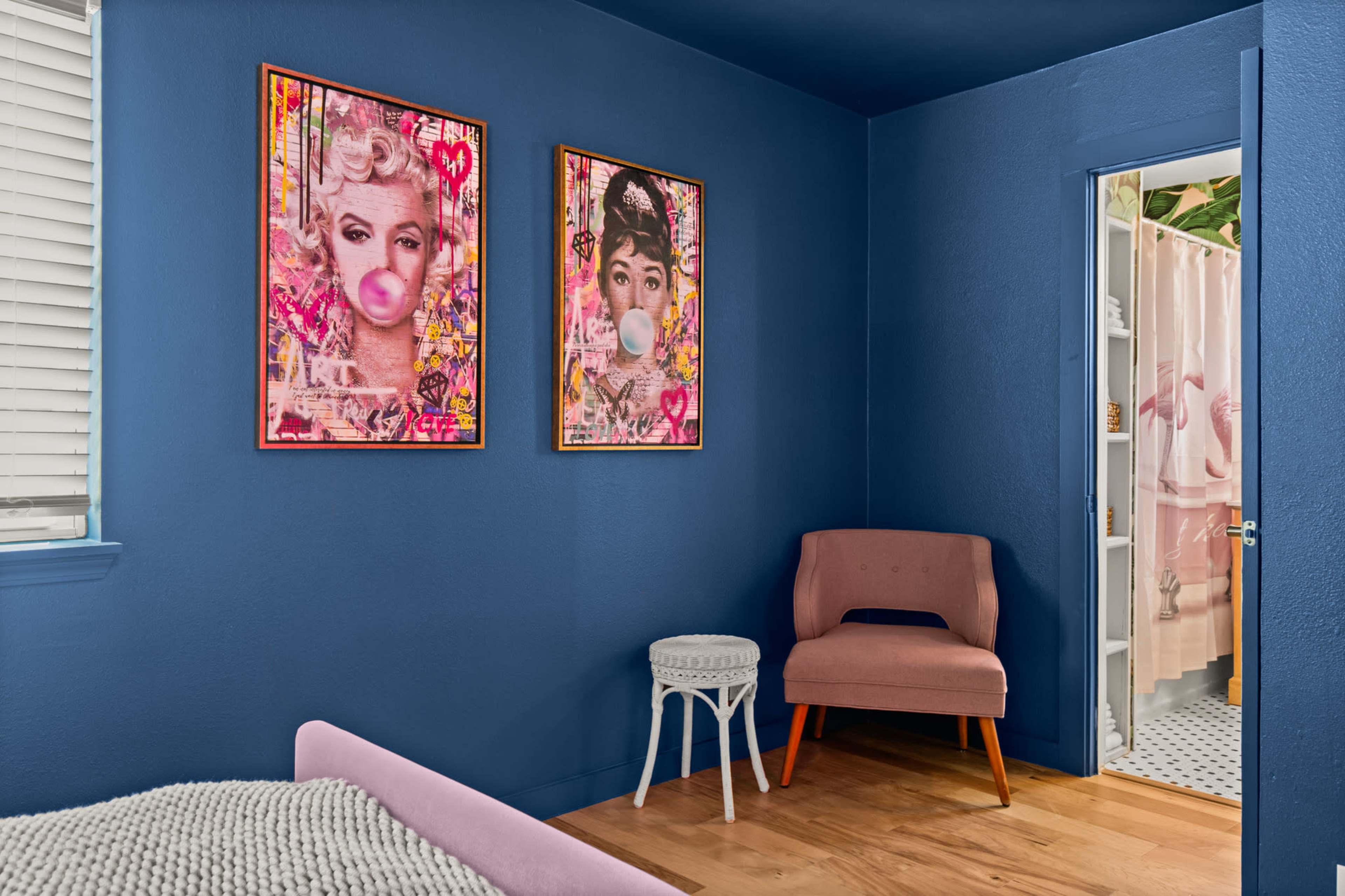 The image shows a corner of a room with blue walls, featuring two colorful art pieces of women on the wall, a pink chair, and a doorway leading to a bathroom with a shower curtain.