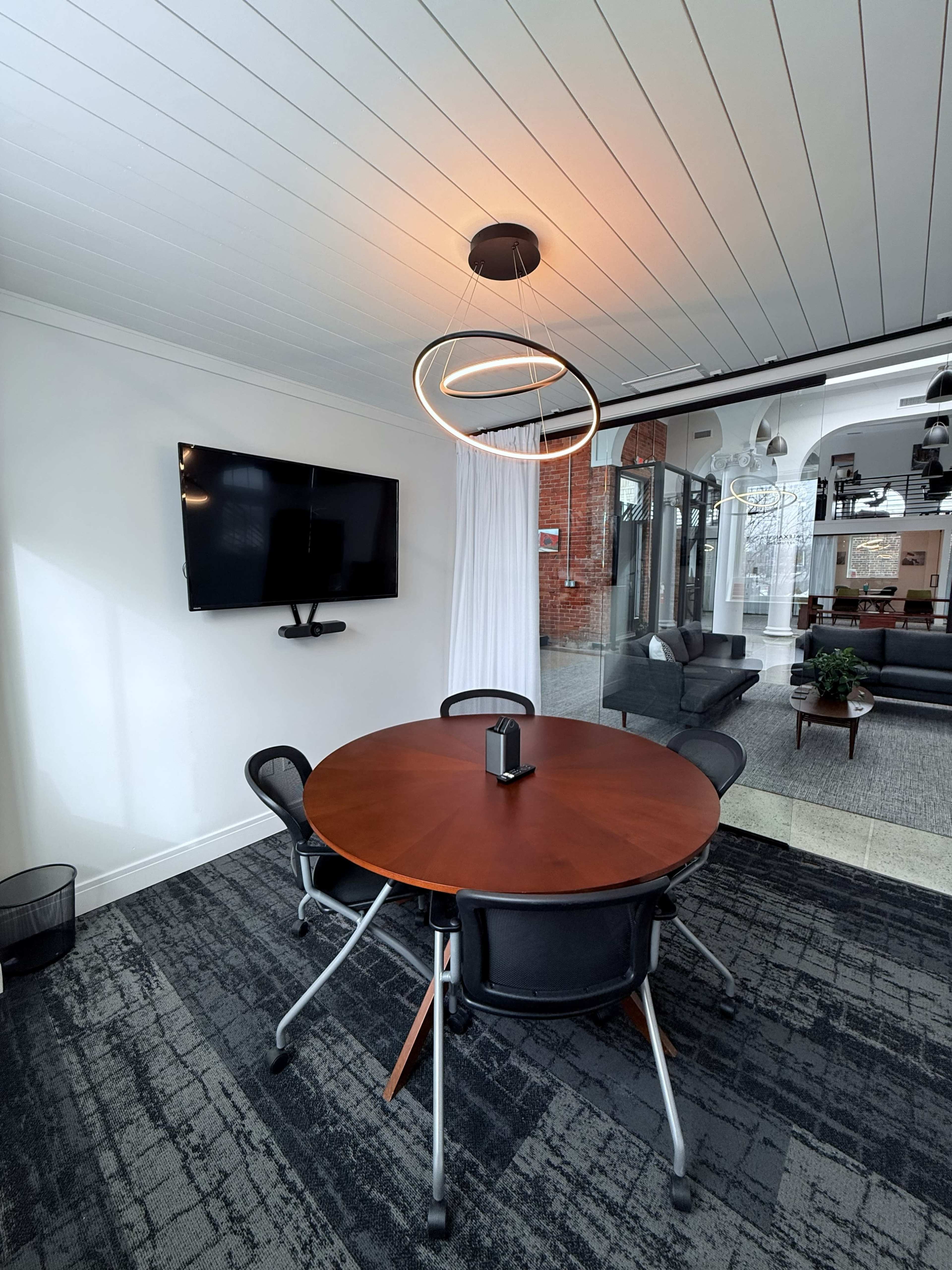 The image shows a modern conference room with a round wooden table, four black chairs, a ceiling light fixture, and a wall-mounted television.