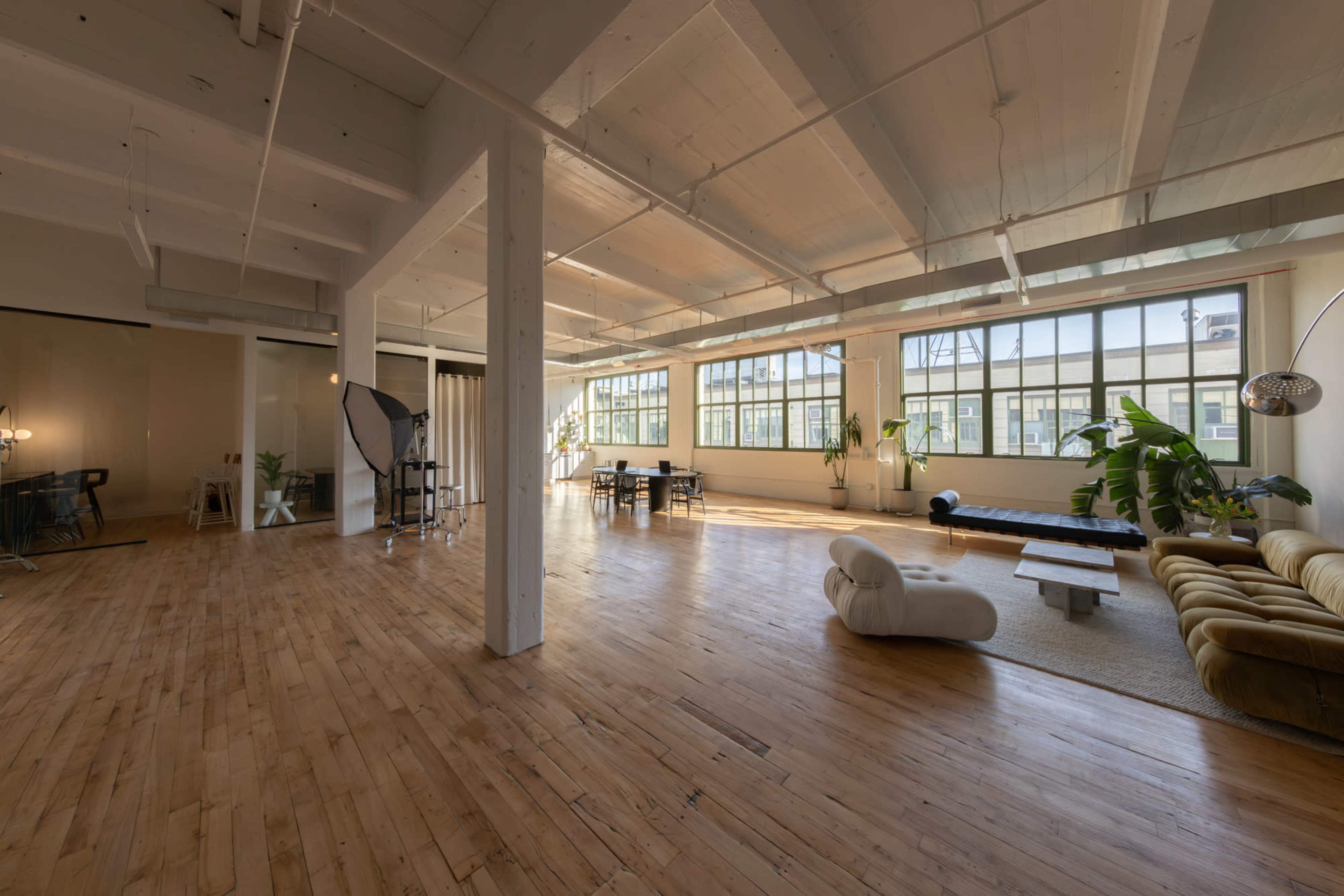 The image shows a spacious, well-lit studio apartment with wooden flooring, large windows, and a mix of modern furniture, including a couch and dining table.