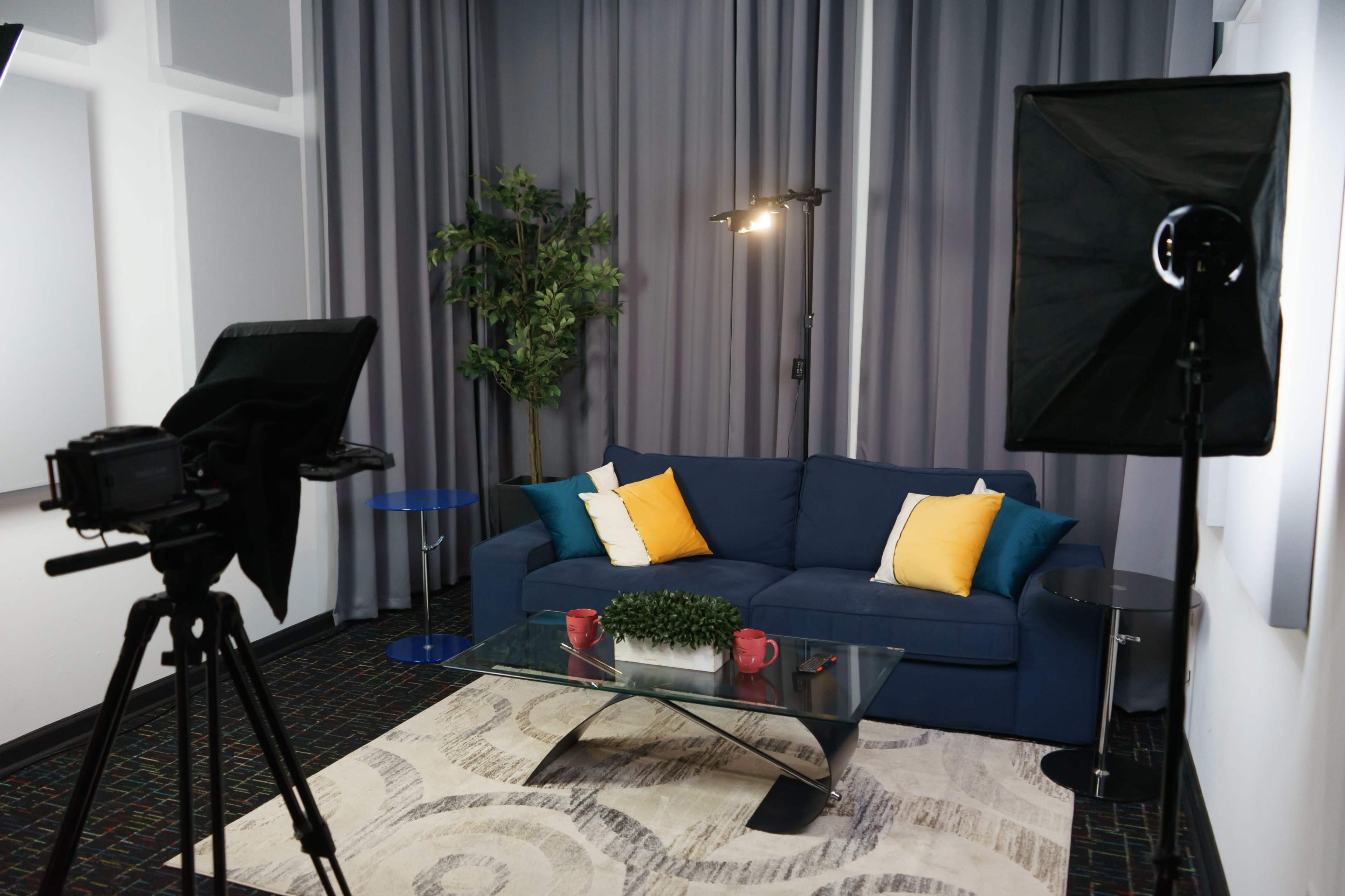 A modern living room setup with a blue sofa, yellow cushions, a glass coffee table, and two lighting fixtures, surrounded by gray curtains and a plant.