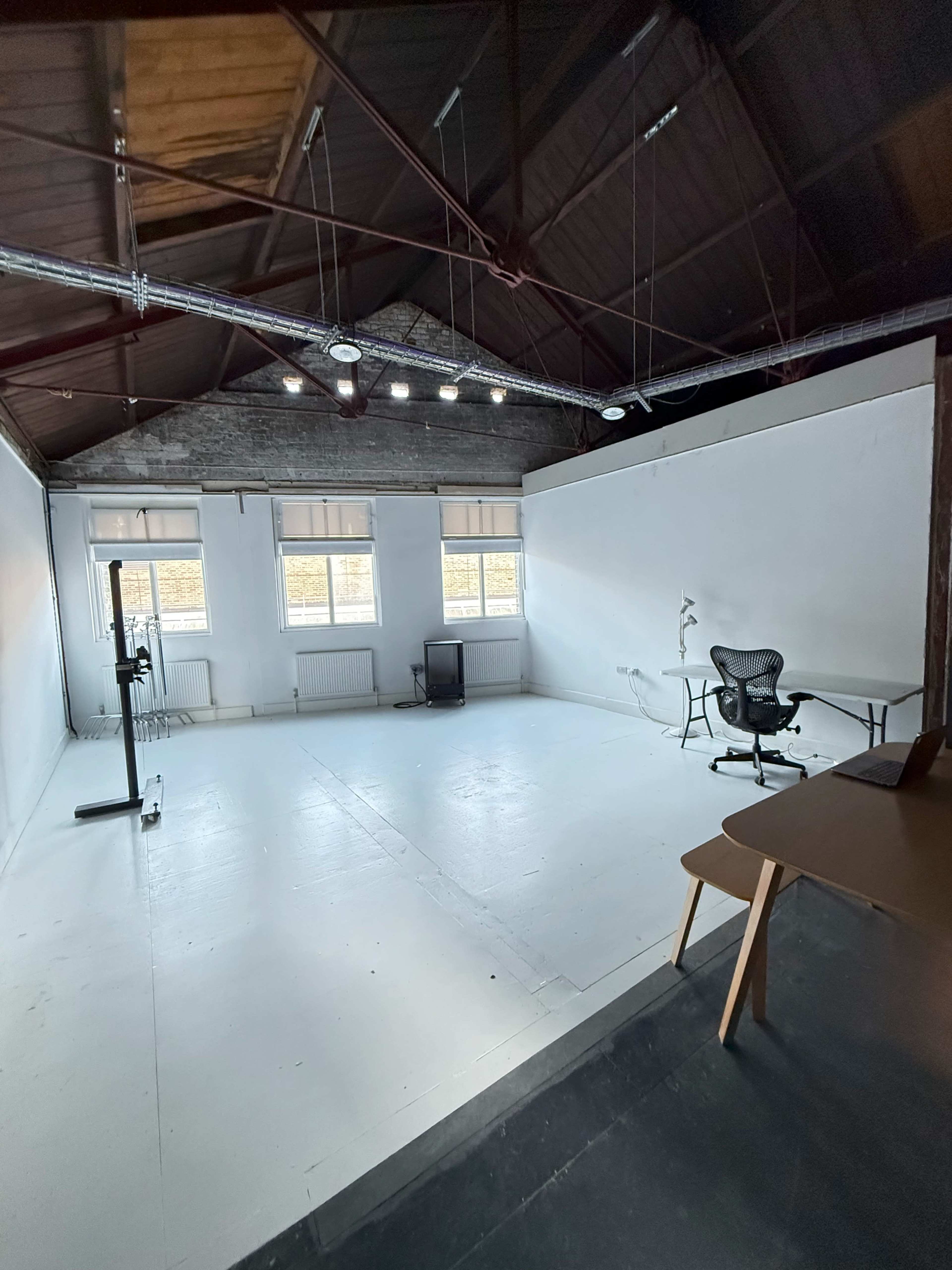 The image shows a spacious, empty studio with large windows, a sleek desk, an office chair, and minimal furniture.