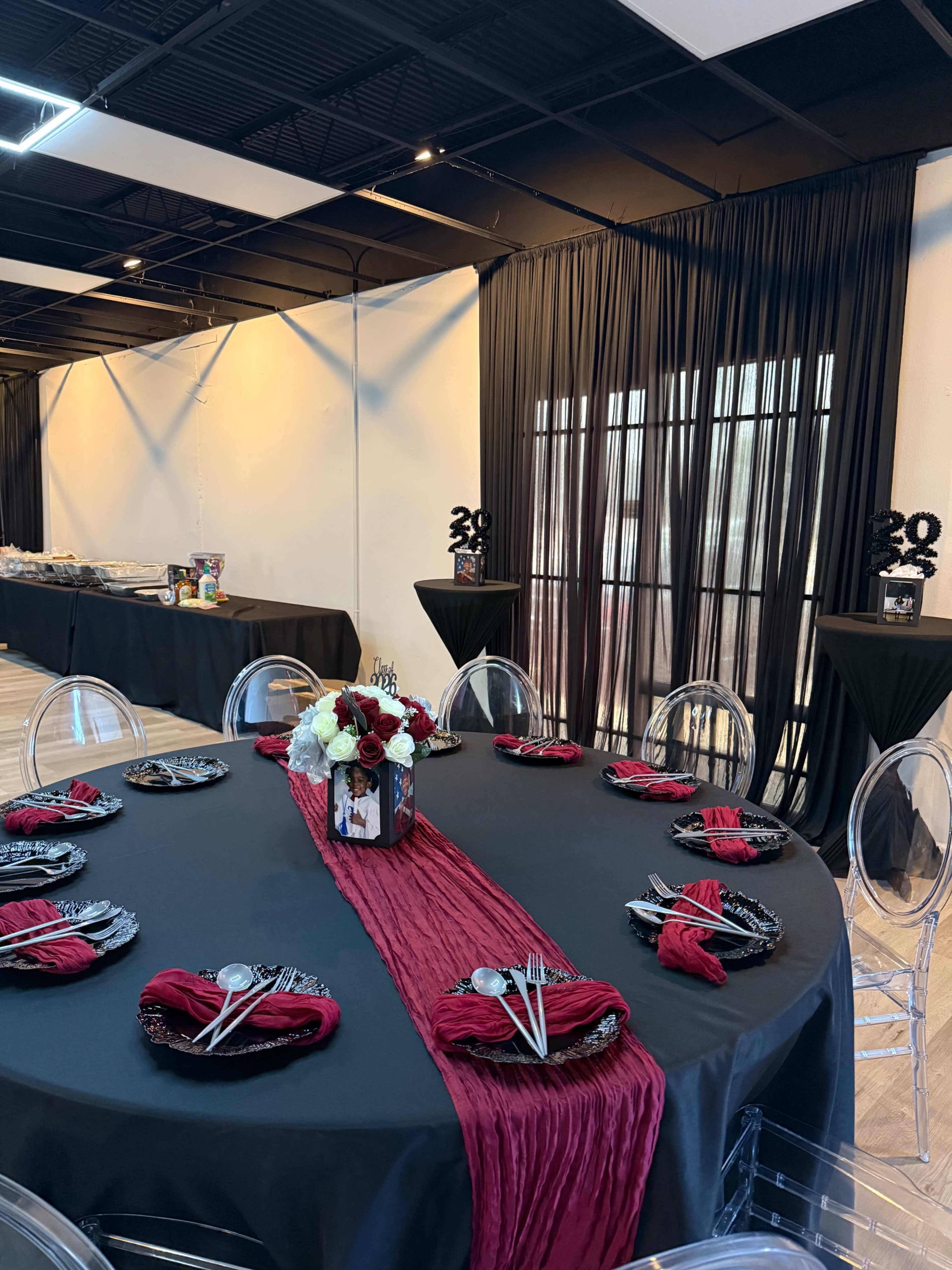 The image shows a decorated event space with a black tablecloth and red accents, featuring a round table set with elegant tableware and floral arrangements, alongside a buffet area in the background.
