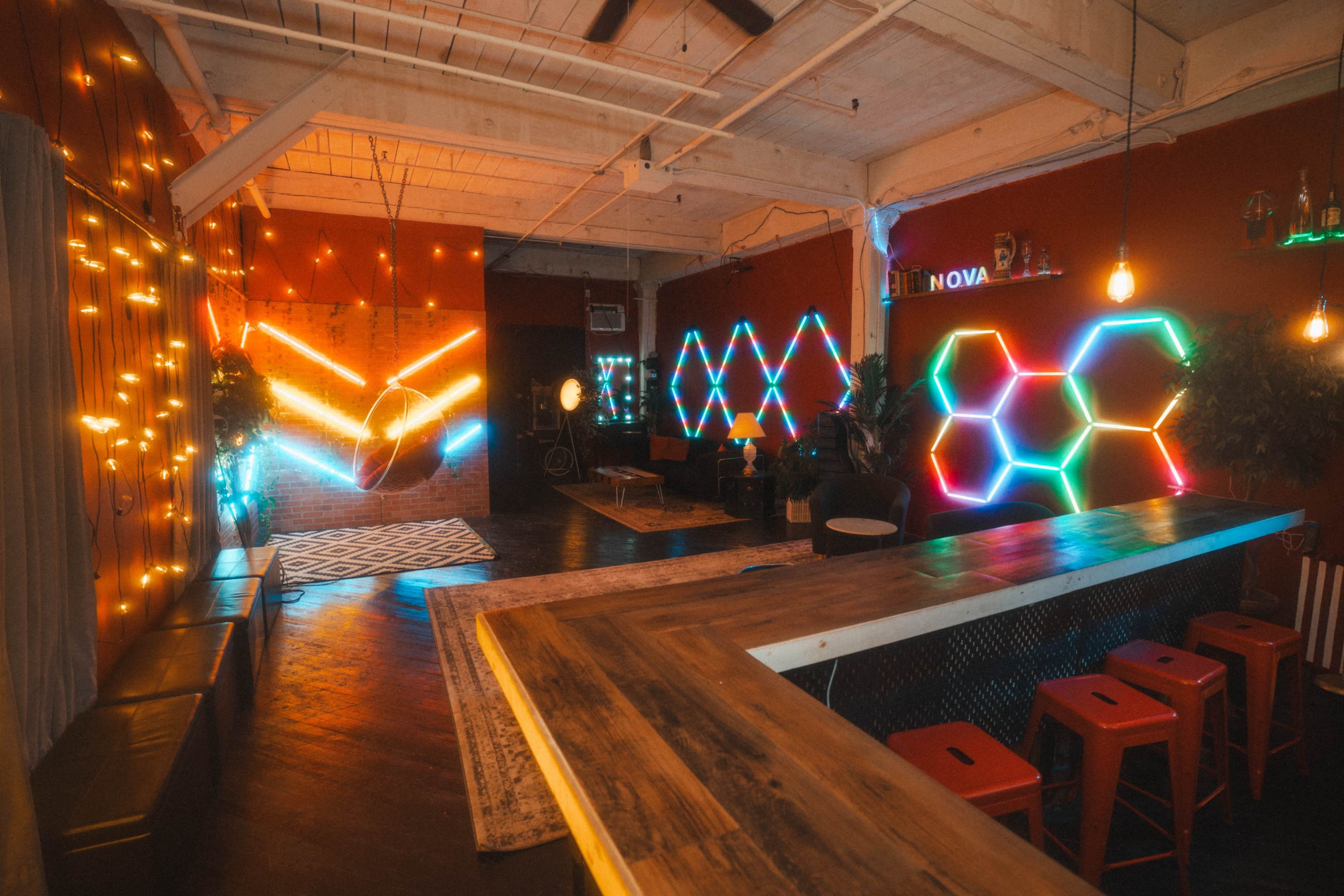 The image shows a modern indoor space with colorful neon lights, lounge seating, and a bar area featuring a wooden counter and red stools.
