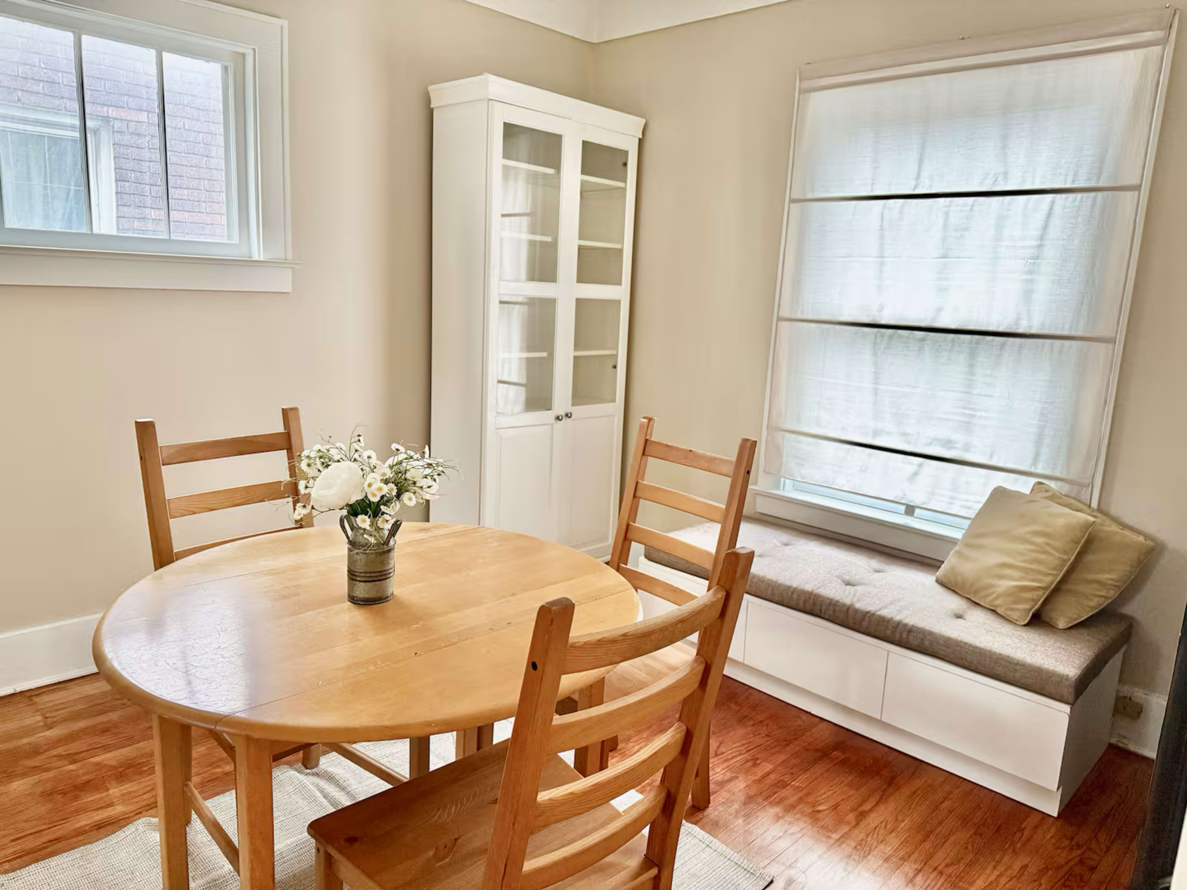 The image shows a small dining area with a round wooden table and four chairs, accompanied by a white cabinet and a window seat beneath a window with partially drawn curtains.