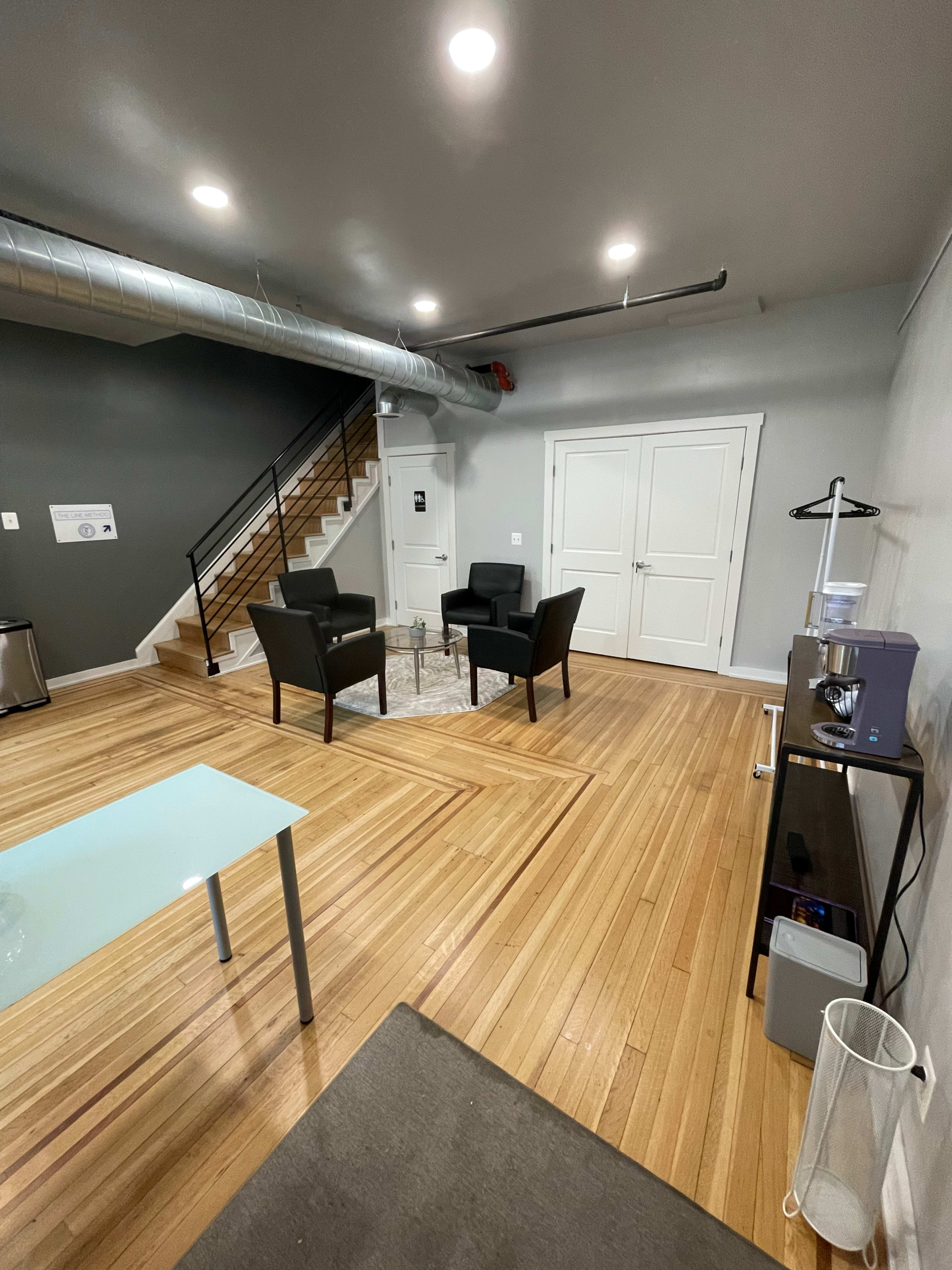 The image shows a modern office space featuring two black armchairs on a rug, a glass table, a staircase, and a cabinet along the wall.