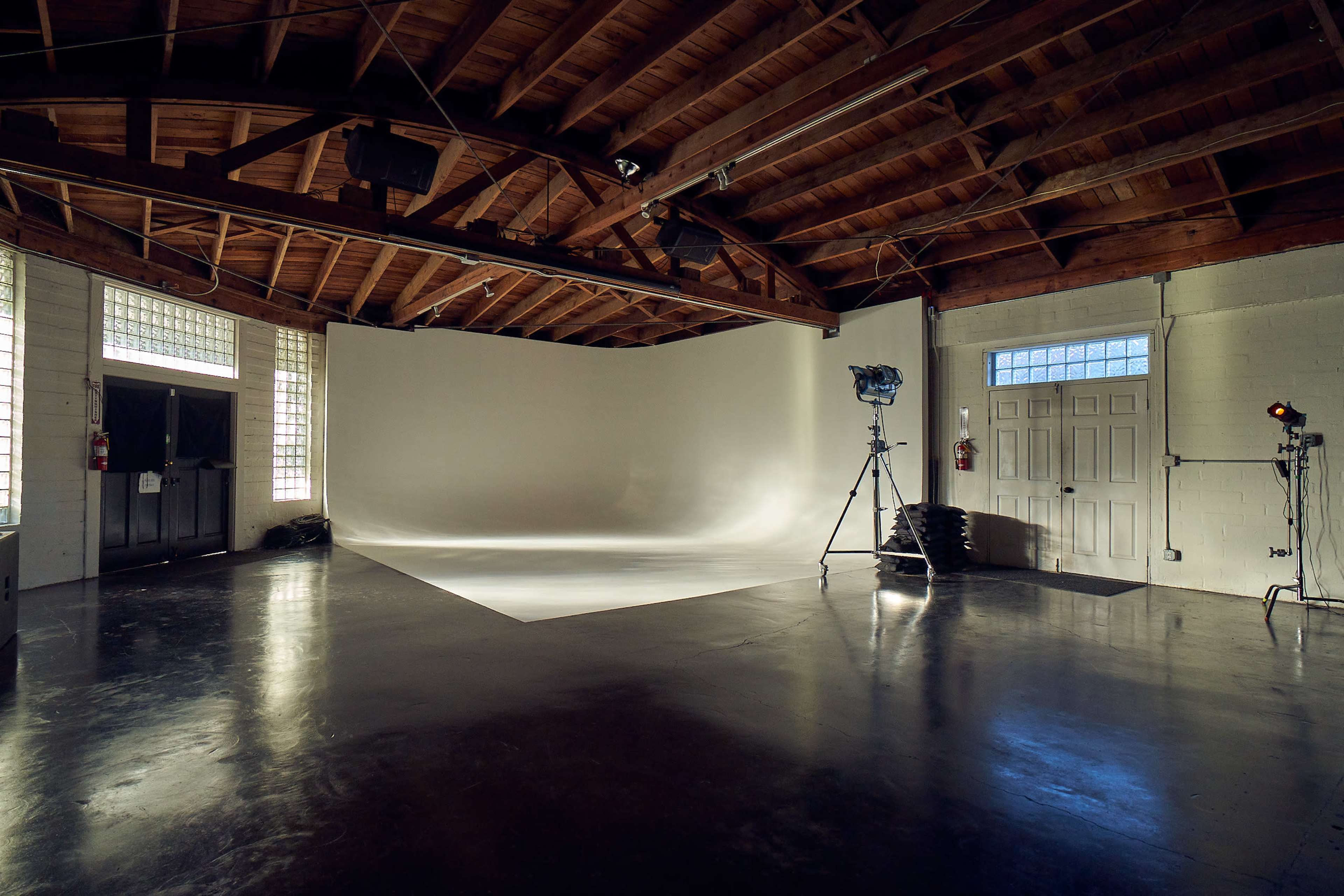 A spacious photography studio features a large seamless backdrop, lighting equipment, and a high ceiling with wooden beams.