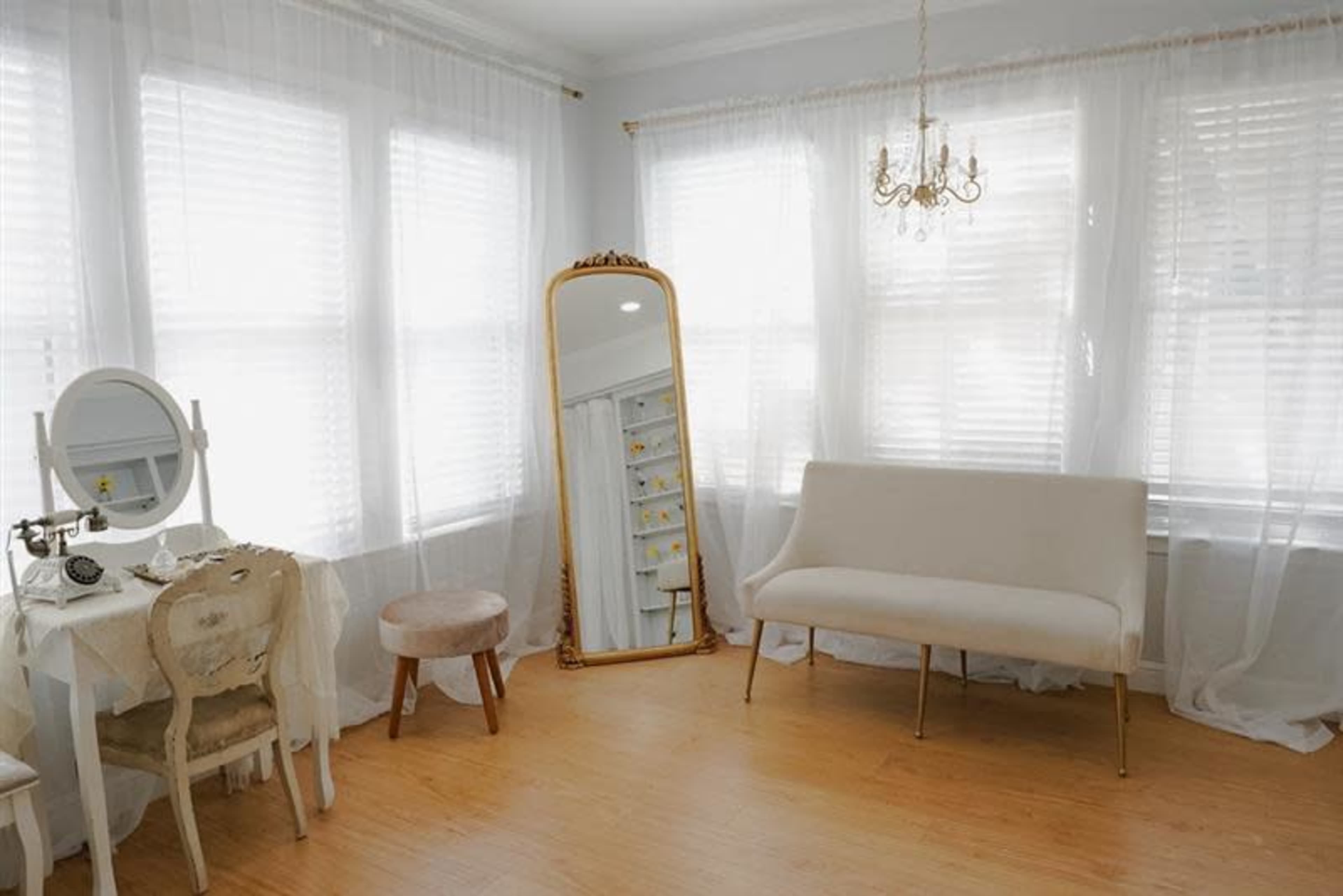 The room features a large mirror, a small vanity table with a stool, and a simple couch, all illuminated by natural light through sheer curtains.