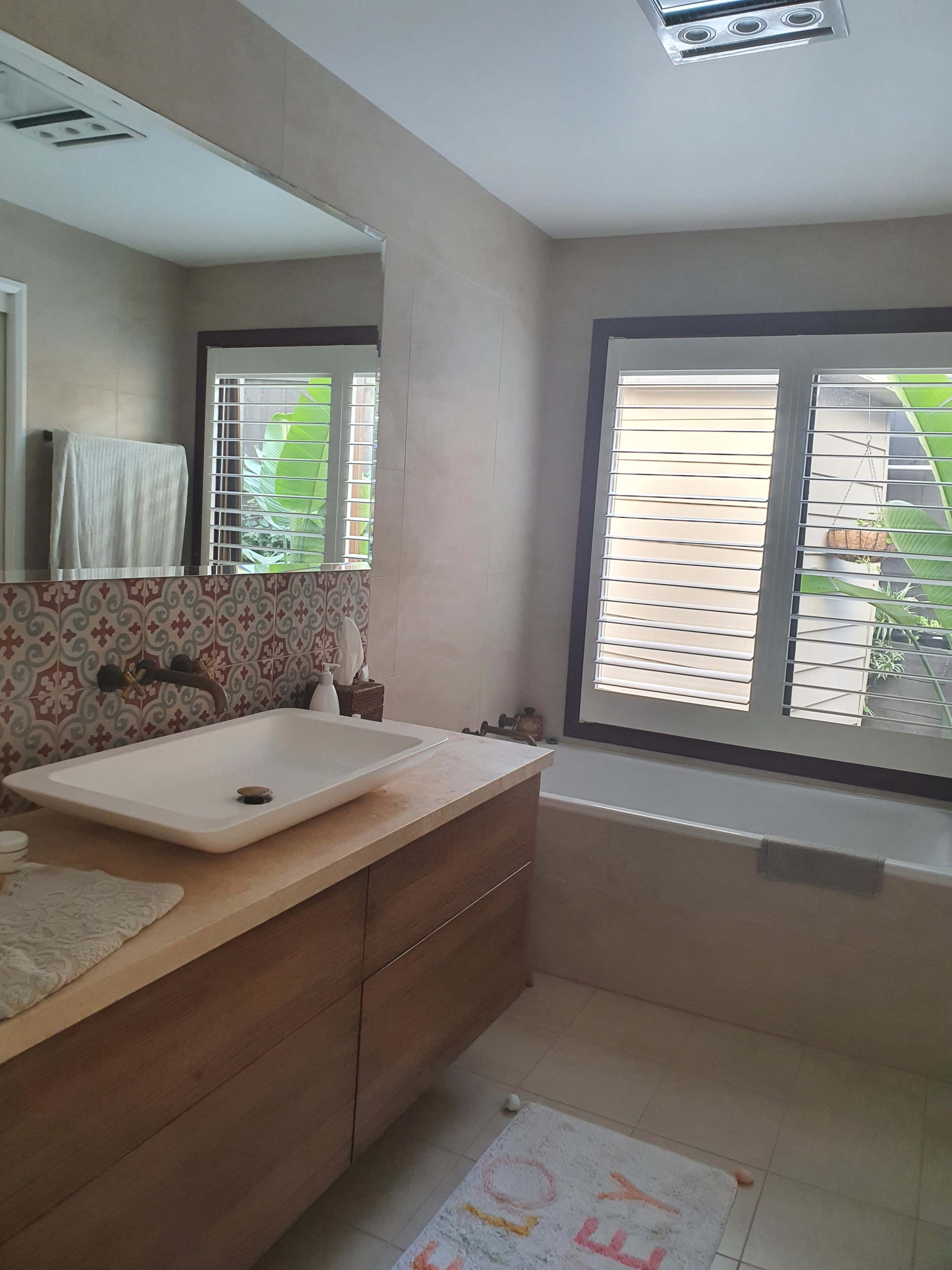 A modern bathroom with a wooden vanity, a large mirror, and a window overlooking greenery.