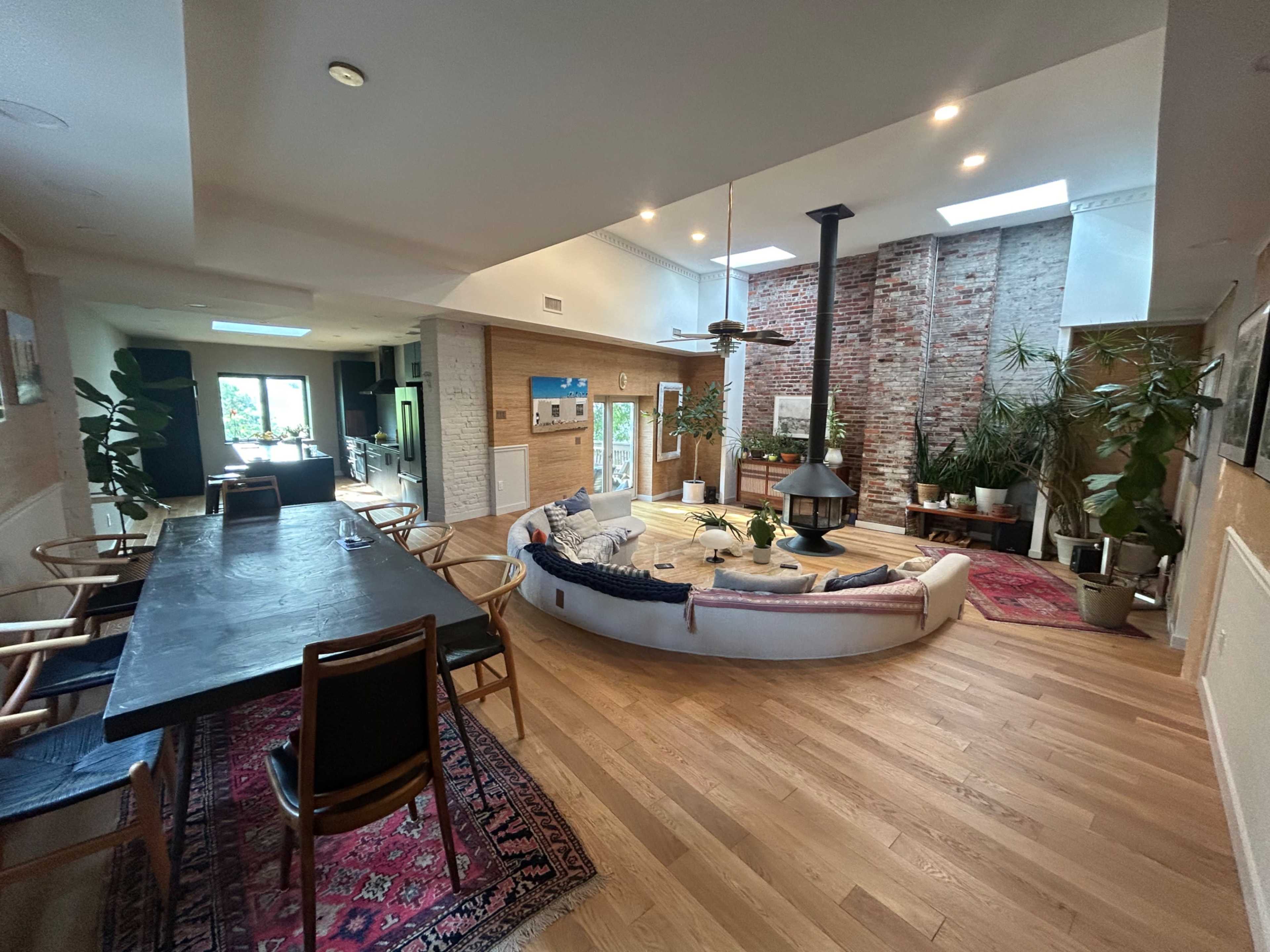 The image shows a modern living space featuring a circular seating area around a central fireplace, with large windows and plants adding to the atmosphere.