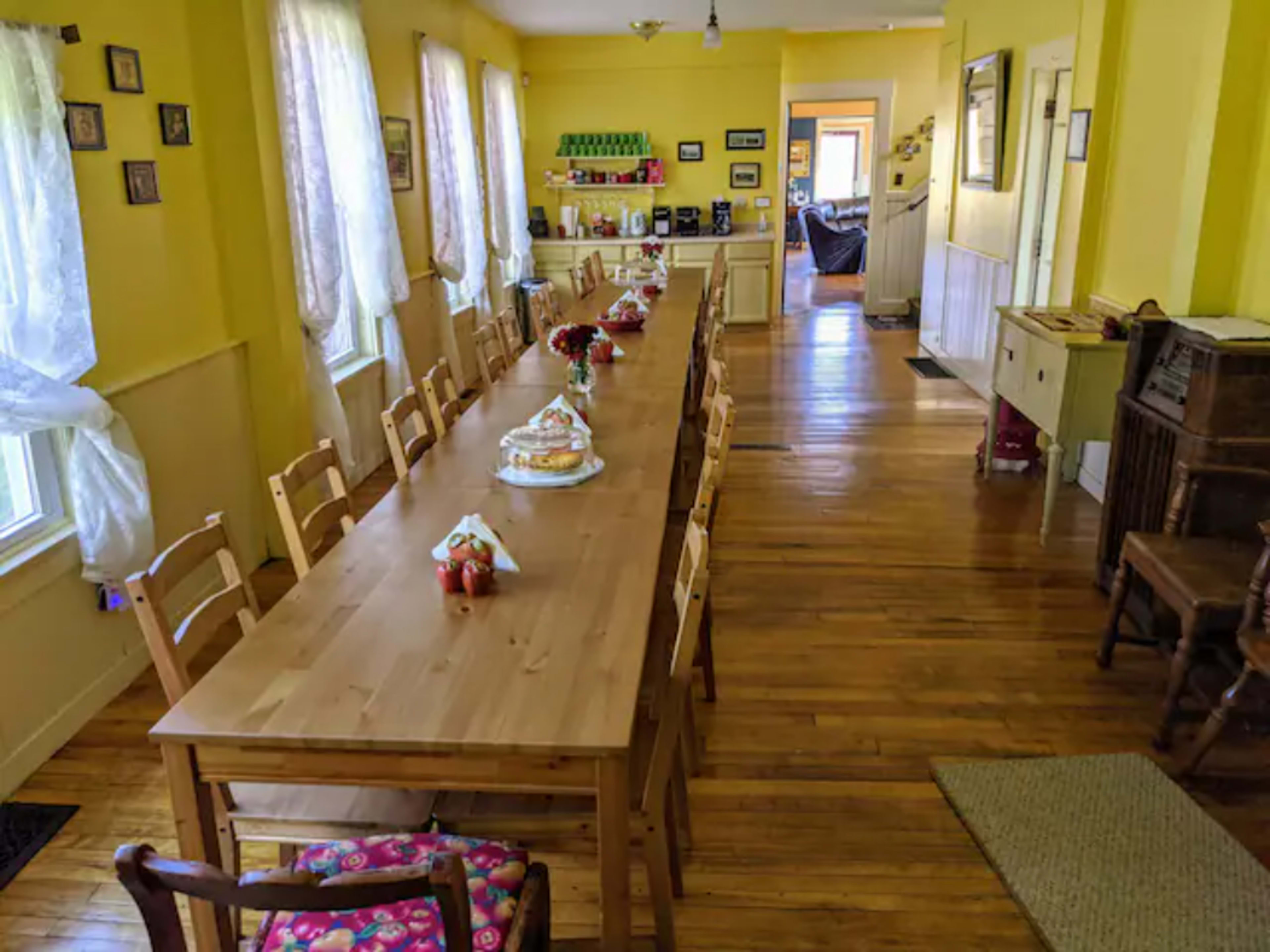 A long wooden dining table is set up in a yellow-walled room with large windows, leading to a visible doorway and another room at the far end.