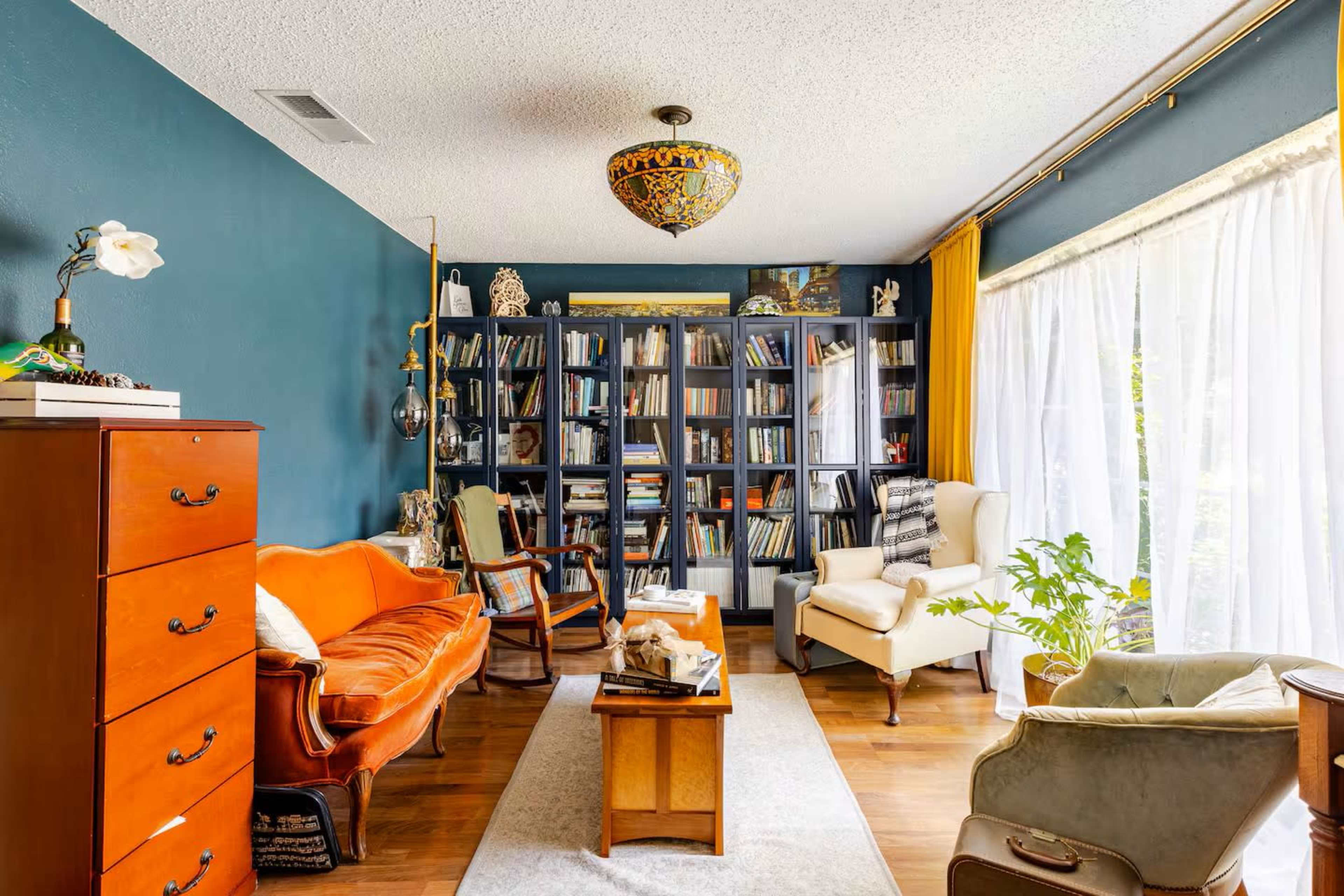 The image shows a cozy living room with a dark blue wall, a collection of bookshelves filled with books, and various pieces of furniture including an orange sofa, an armchair, and a small coffee table.