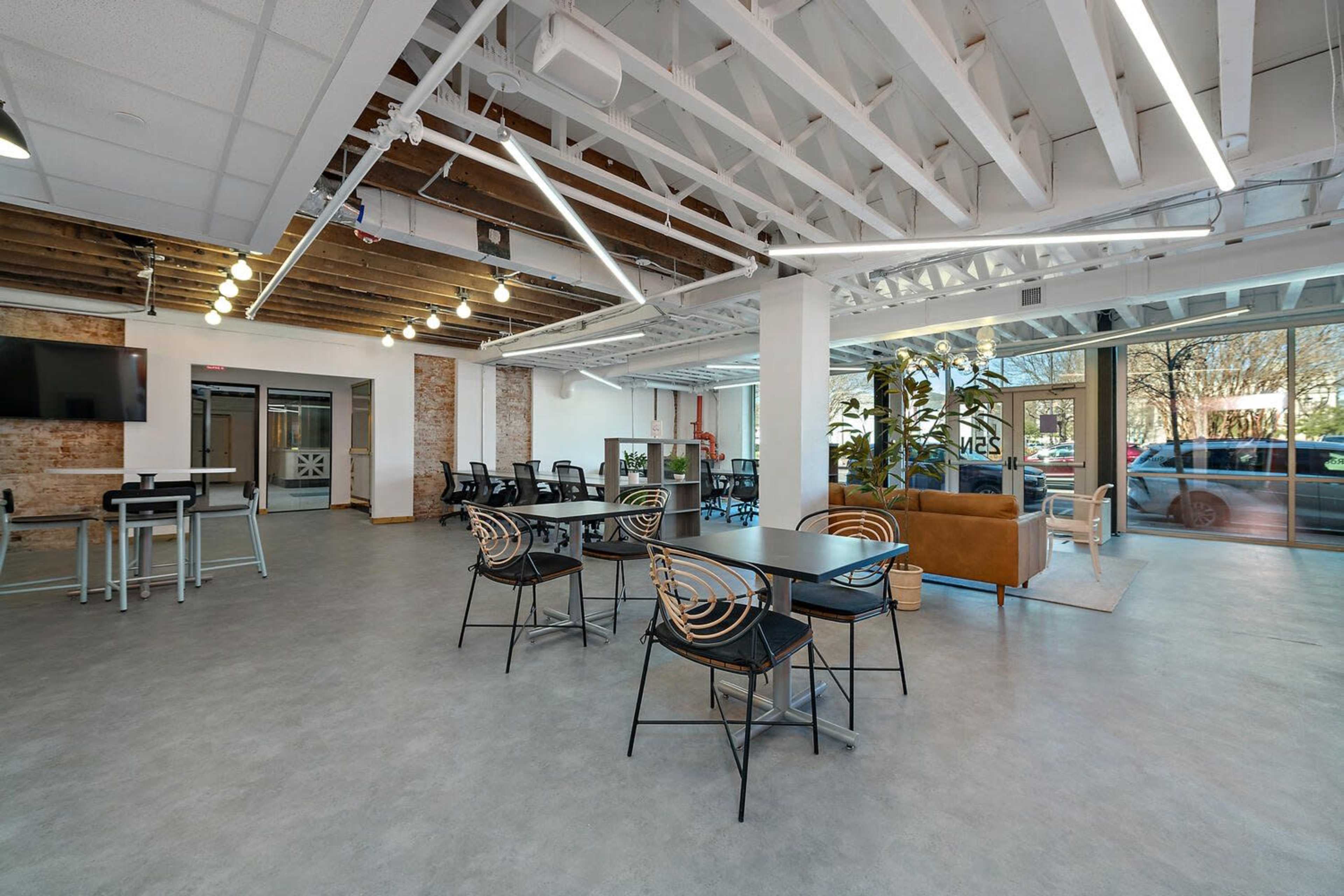 The image shows a modern office space with exposed beams, multiple tables, seating areas, and large windows allowing natural light to enter.