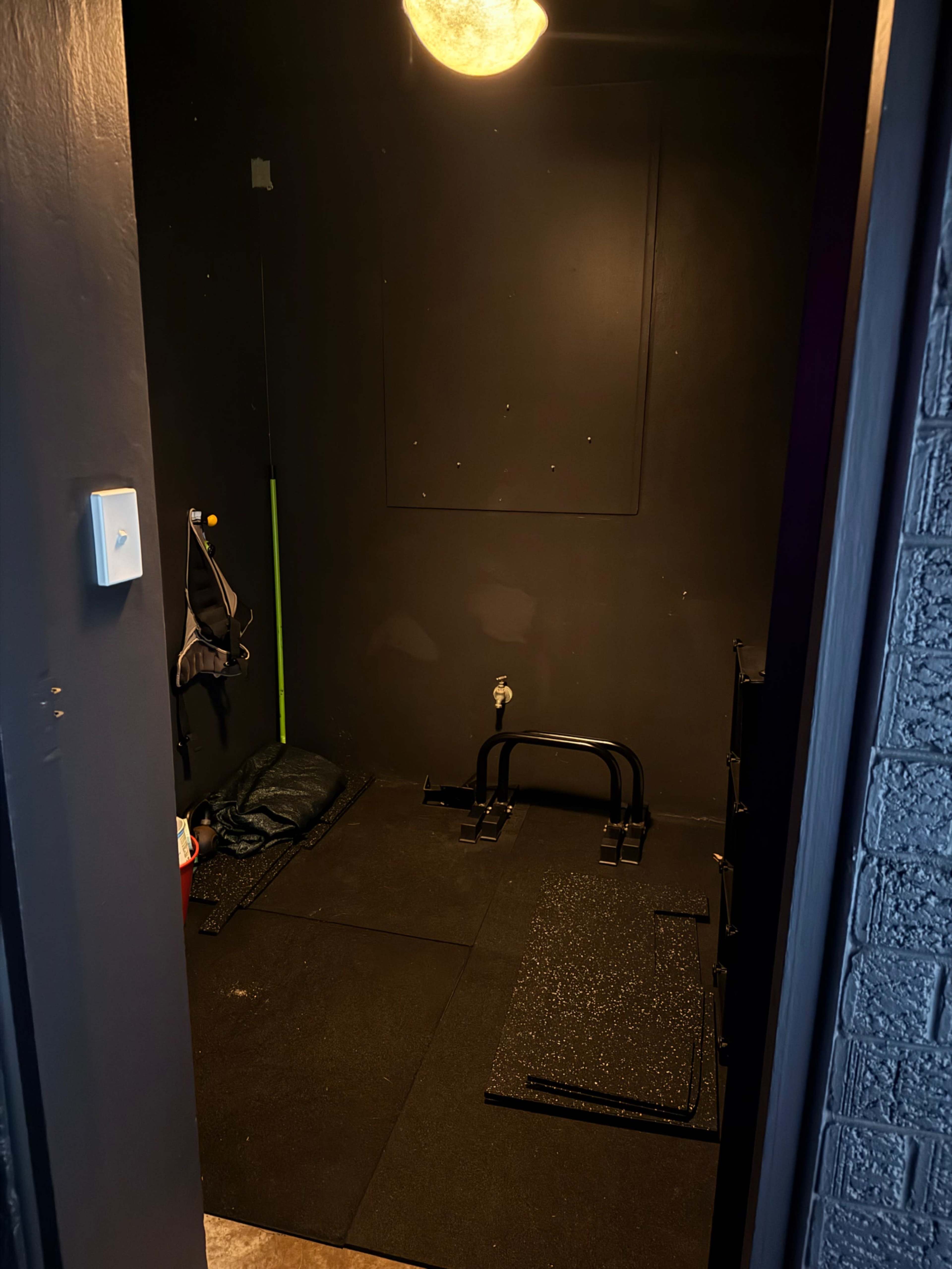 The image shows a dimly lit room with dark walls, equipped with exercise mats and a pull-up bar on the floor.