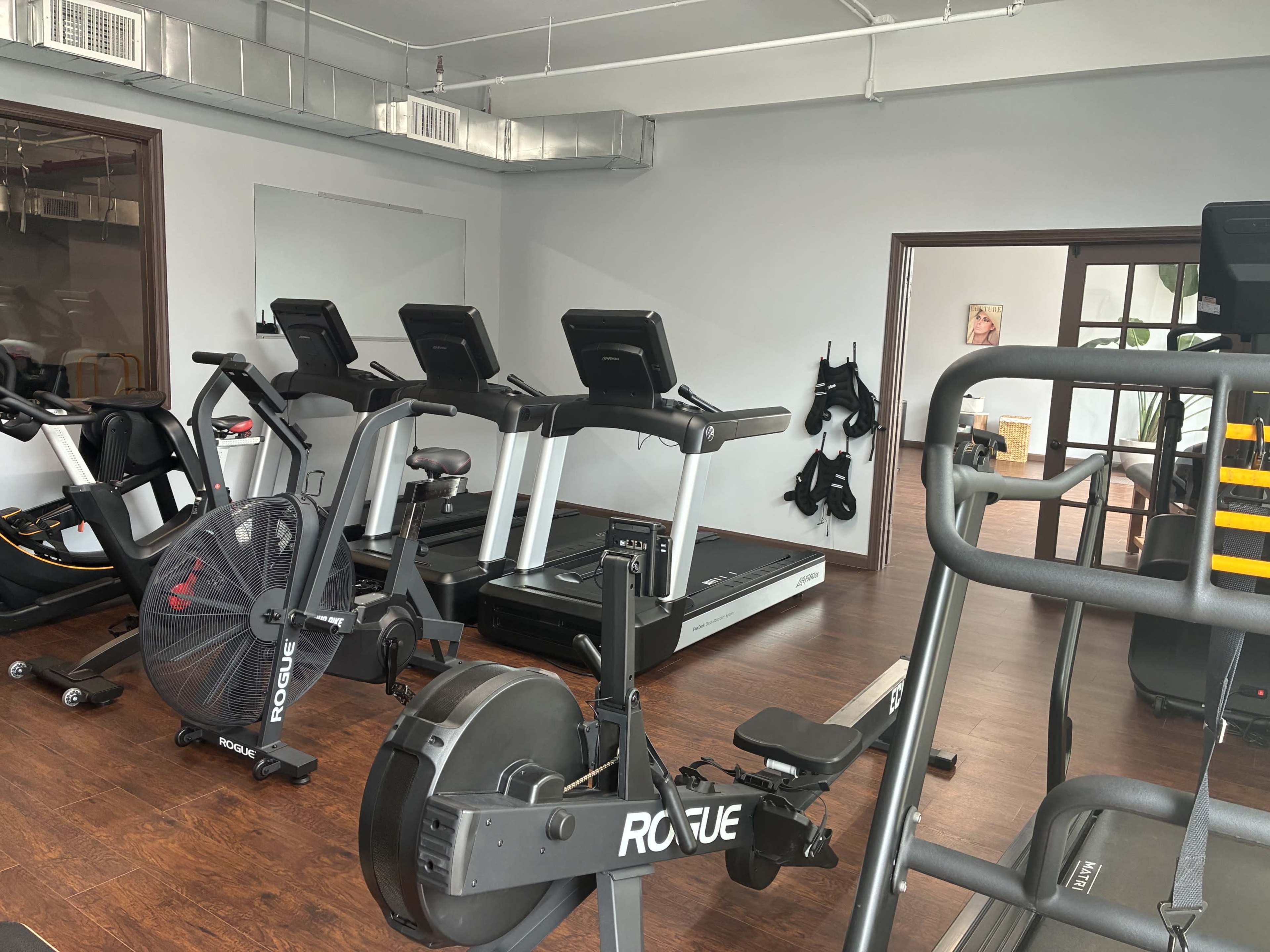 The image shows a small gym with various exercise equipment, including treadmills, a rowing machine, and an air bike, set against a minimalist interior.