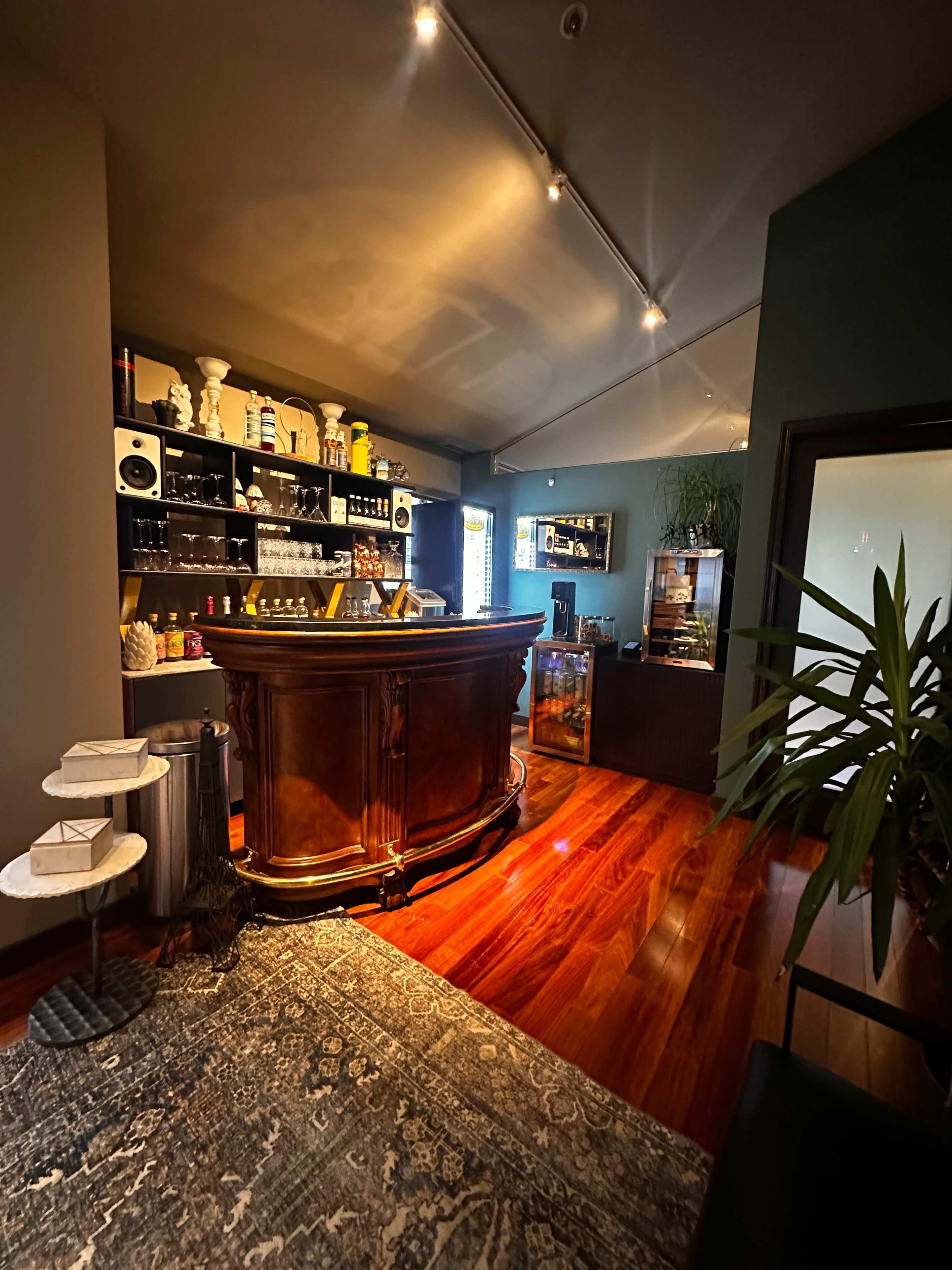 The image shows a sleek bar area with a dark wooden counter, shelves filled with various bottles, and polished hardwood flooring.