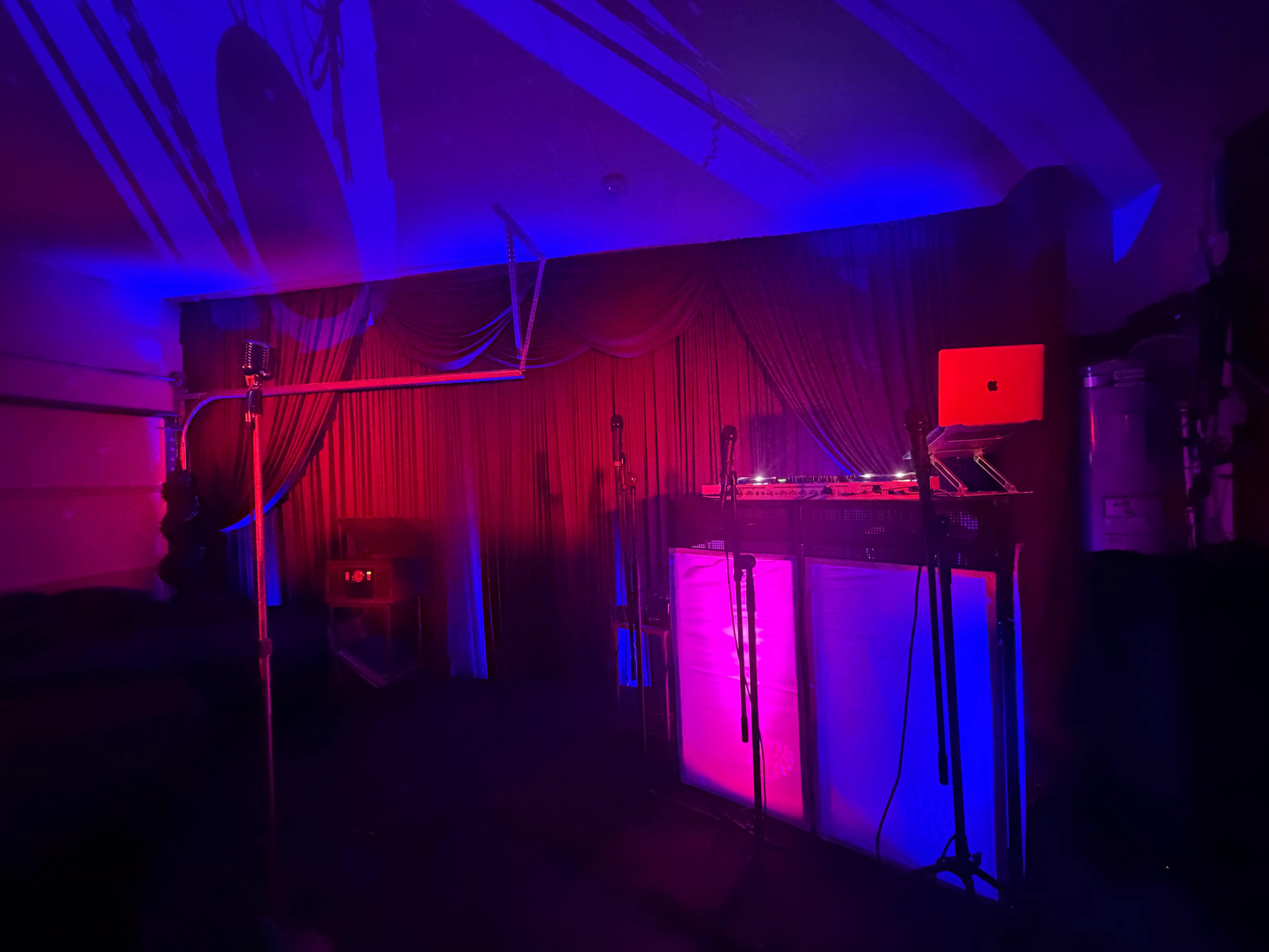 A dimly lit room features a DJ setup with a laptop and microphones against a backdrop of draped red curtains, illuminated by blue and purple lights.