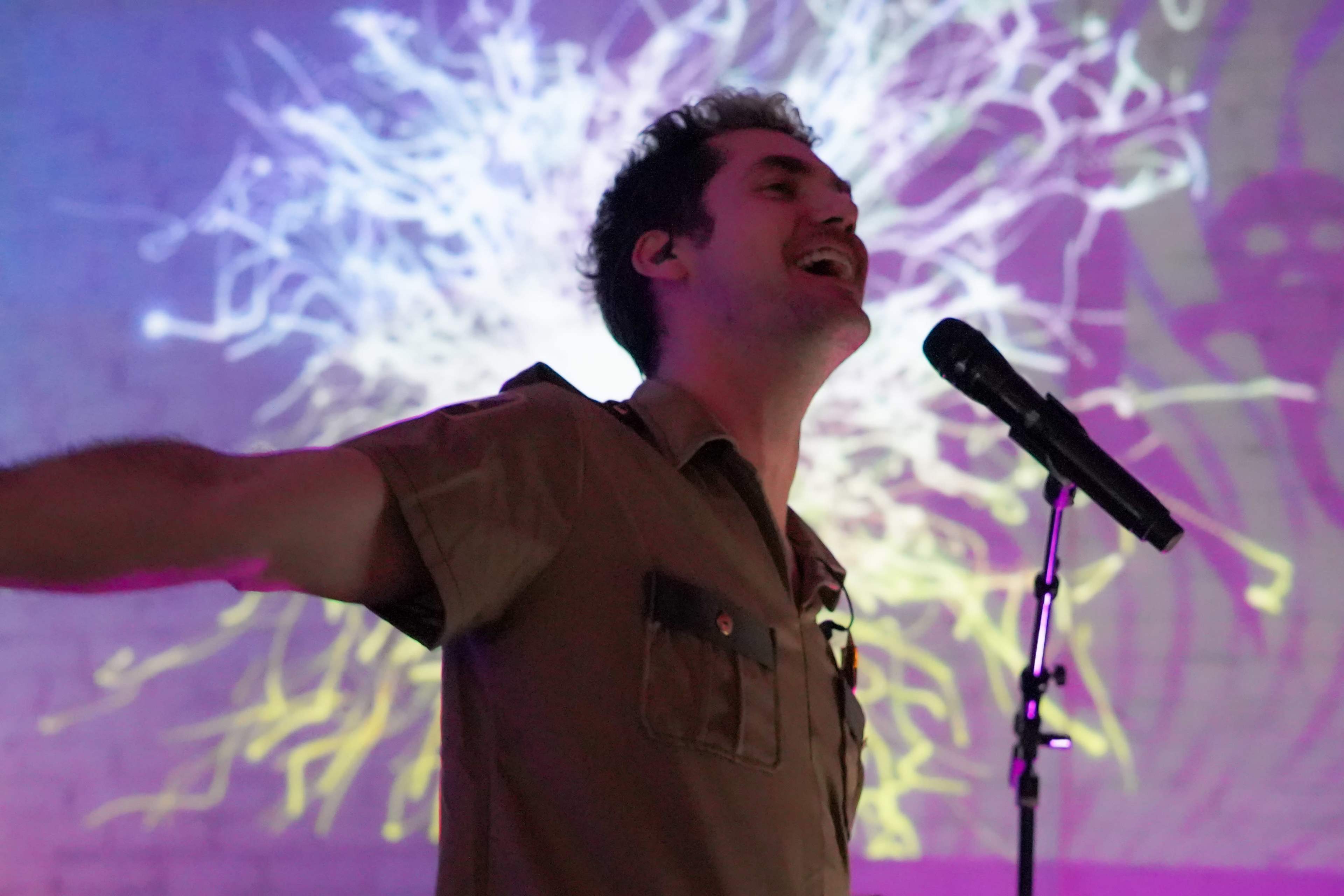 A performer with arms outstretched sings into a microphone against a colorful, abstract background.