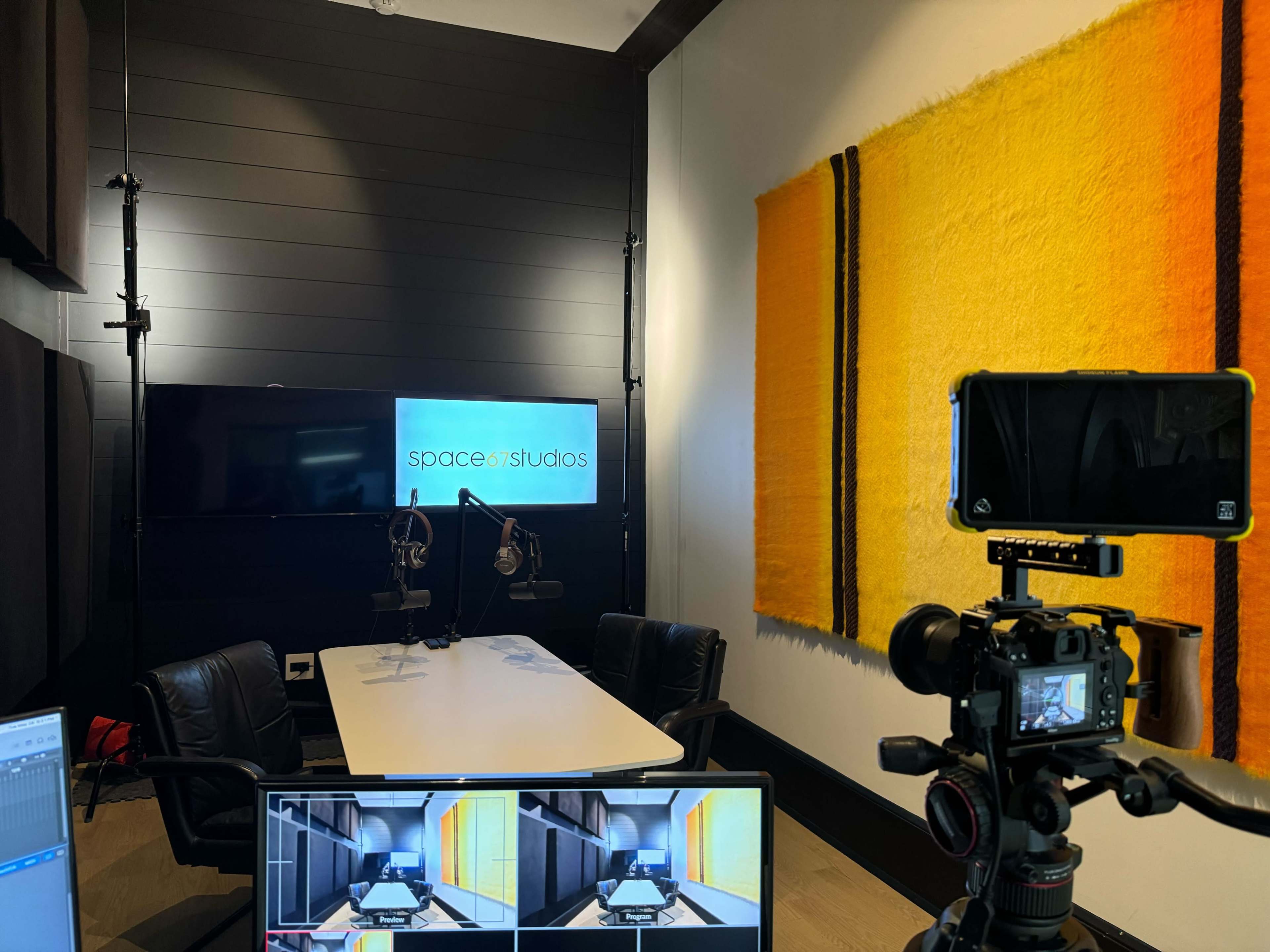 A recording studio with a large screen displaying "space7studios," two microphones on a table, and a camera set up facing the table.