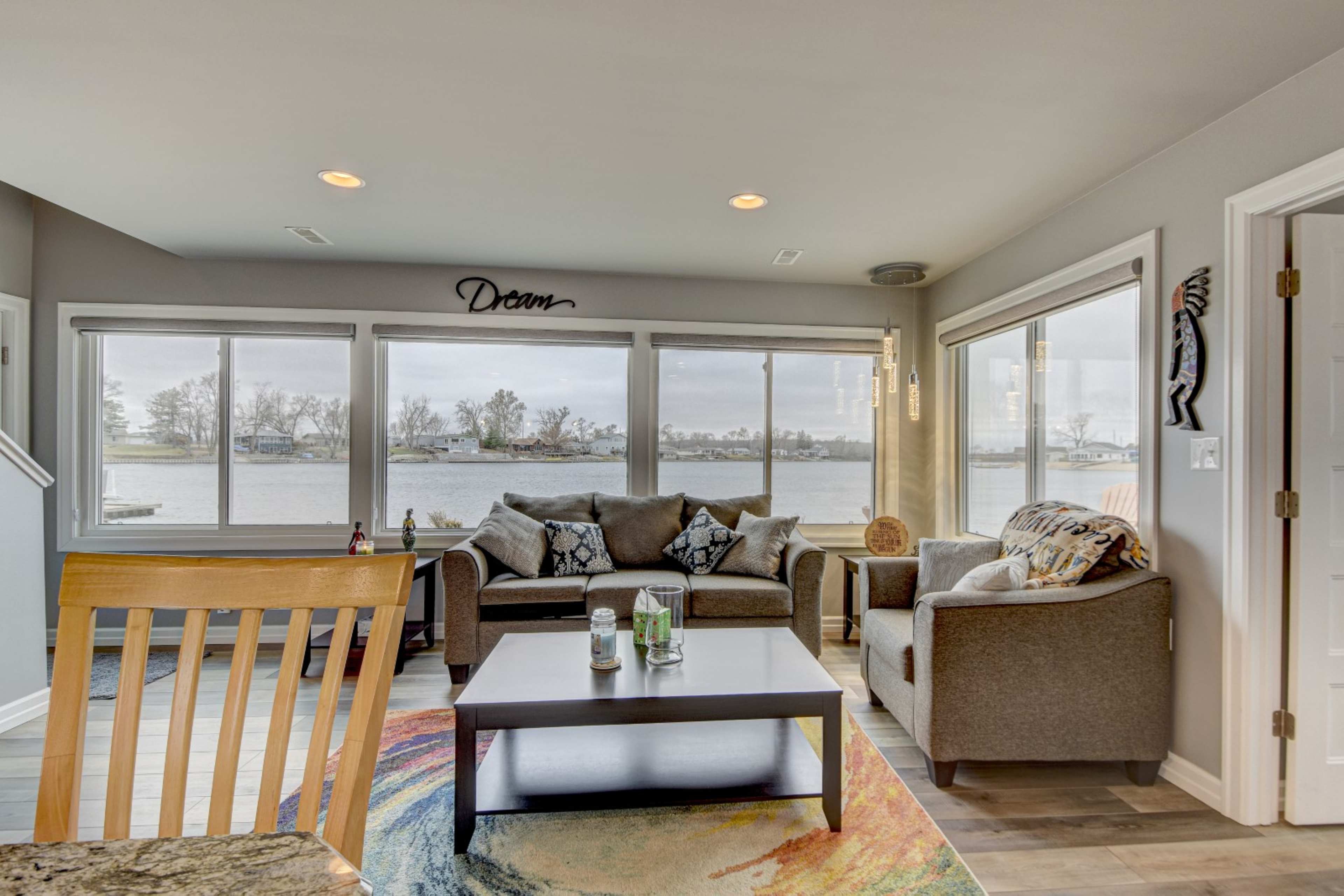 A cozy living room features a gray sectional sofa and chairs facing large windows overlooking a body of water.