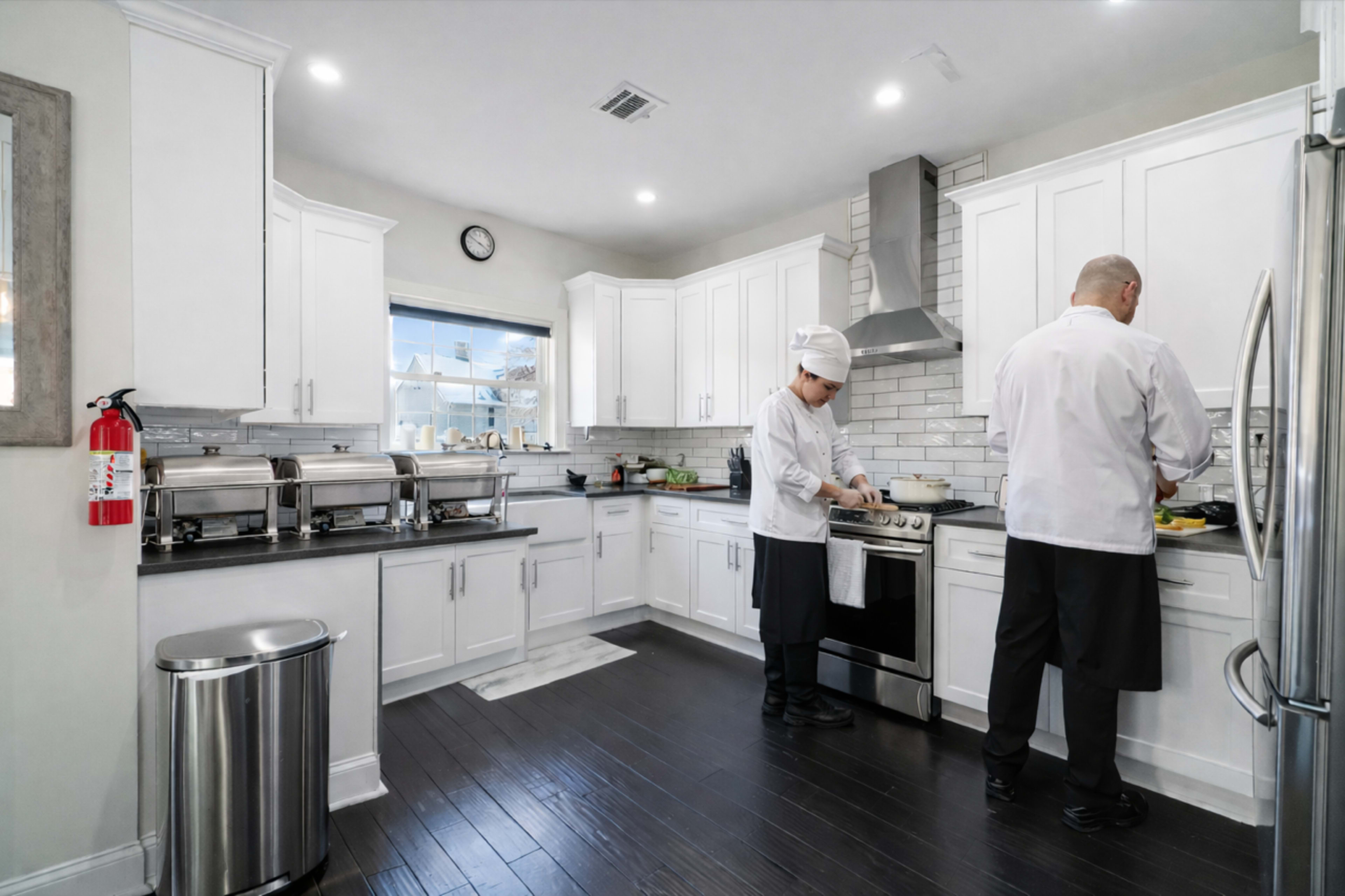 Two chefs in white uniforms prepare food in a modern kitchen equipped with stainless steel appliances and white cabinetry.
