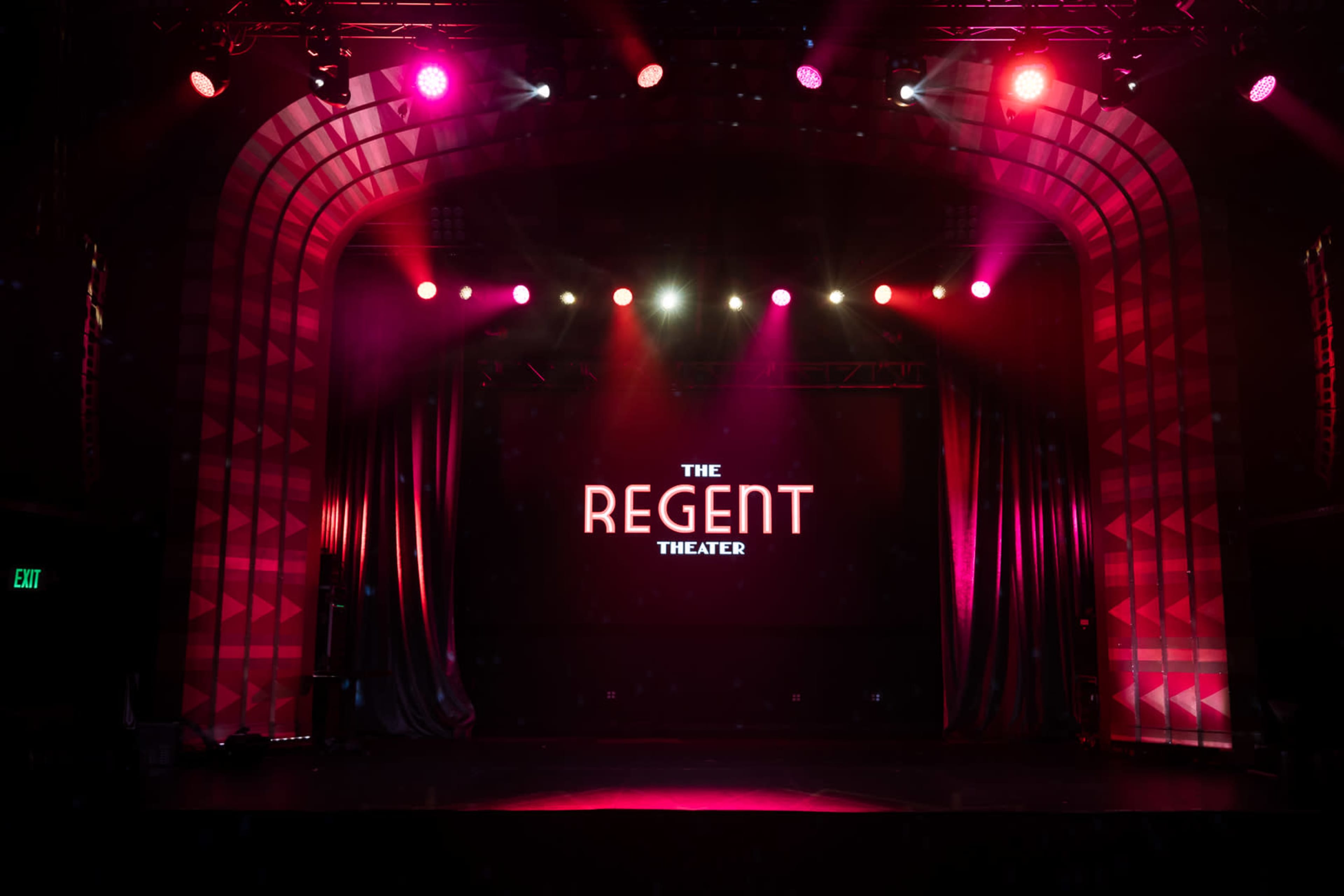 The image shows the interior of the Regent Theater with a lit stage and a large sign displaying "THE REGENT THEATER" against a dark background.