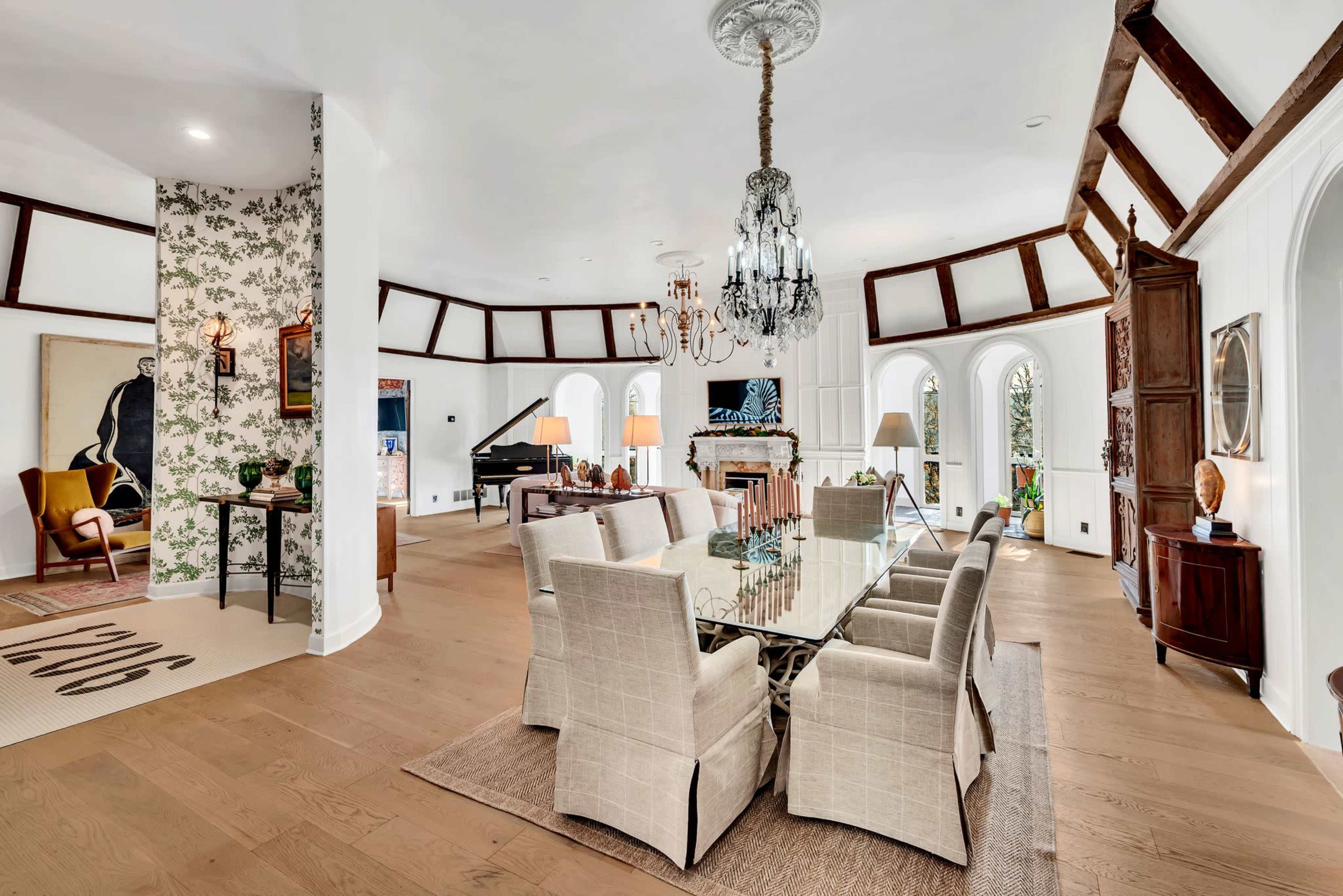 The image depicts an elegantly decorated dining room featuring a glass dining table surrounded by beige chairs, with a piano visible in the background and large windows letting in natural light.