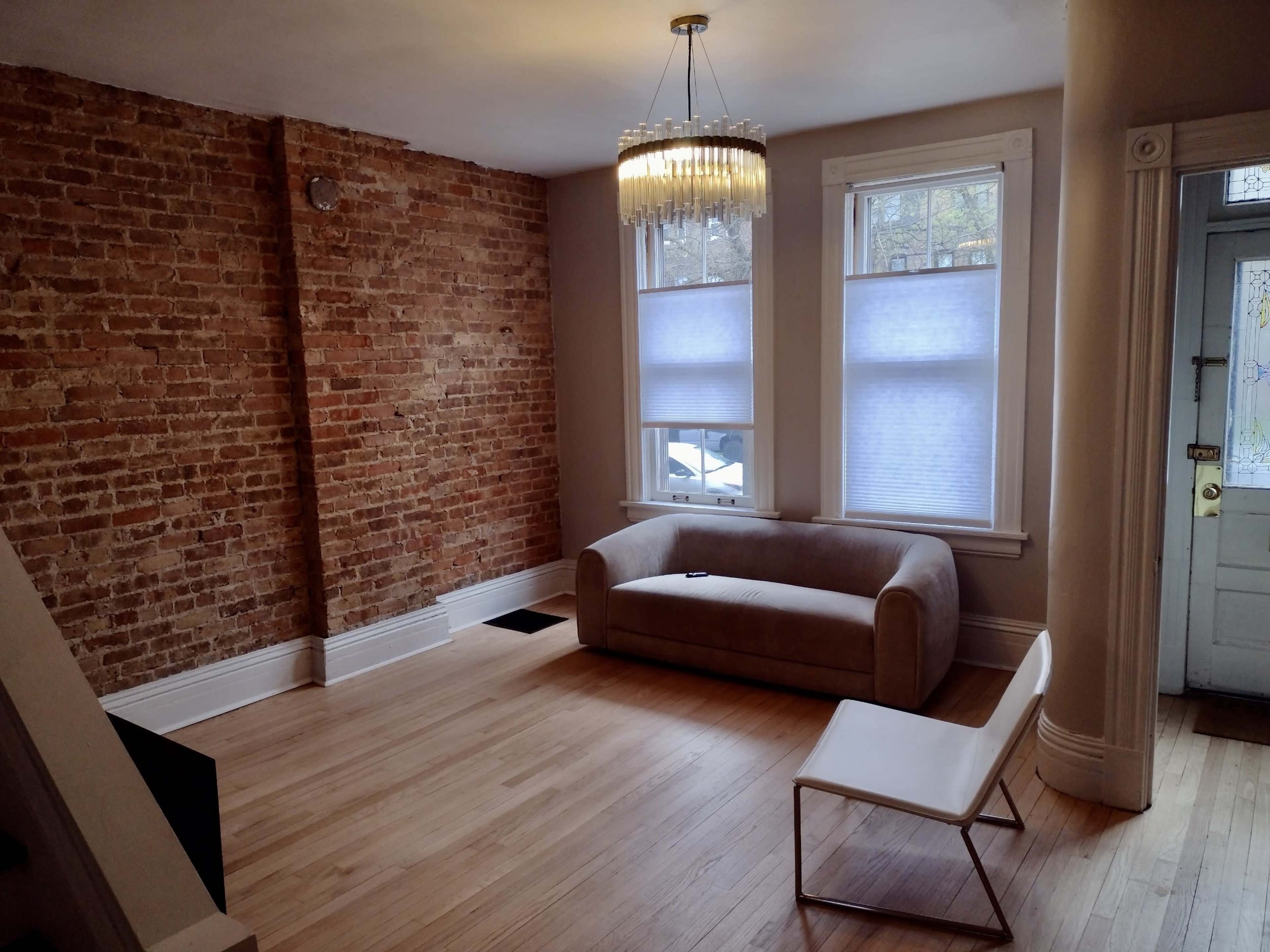 A living room with exposed brick walls, a light-colored sofa, a modern chair, and large windows with shades.