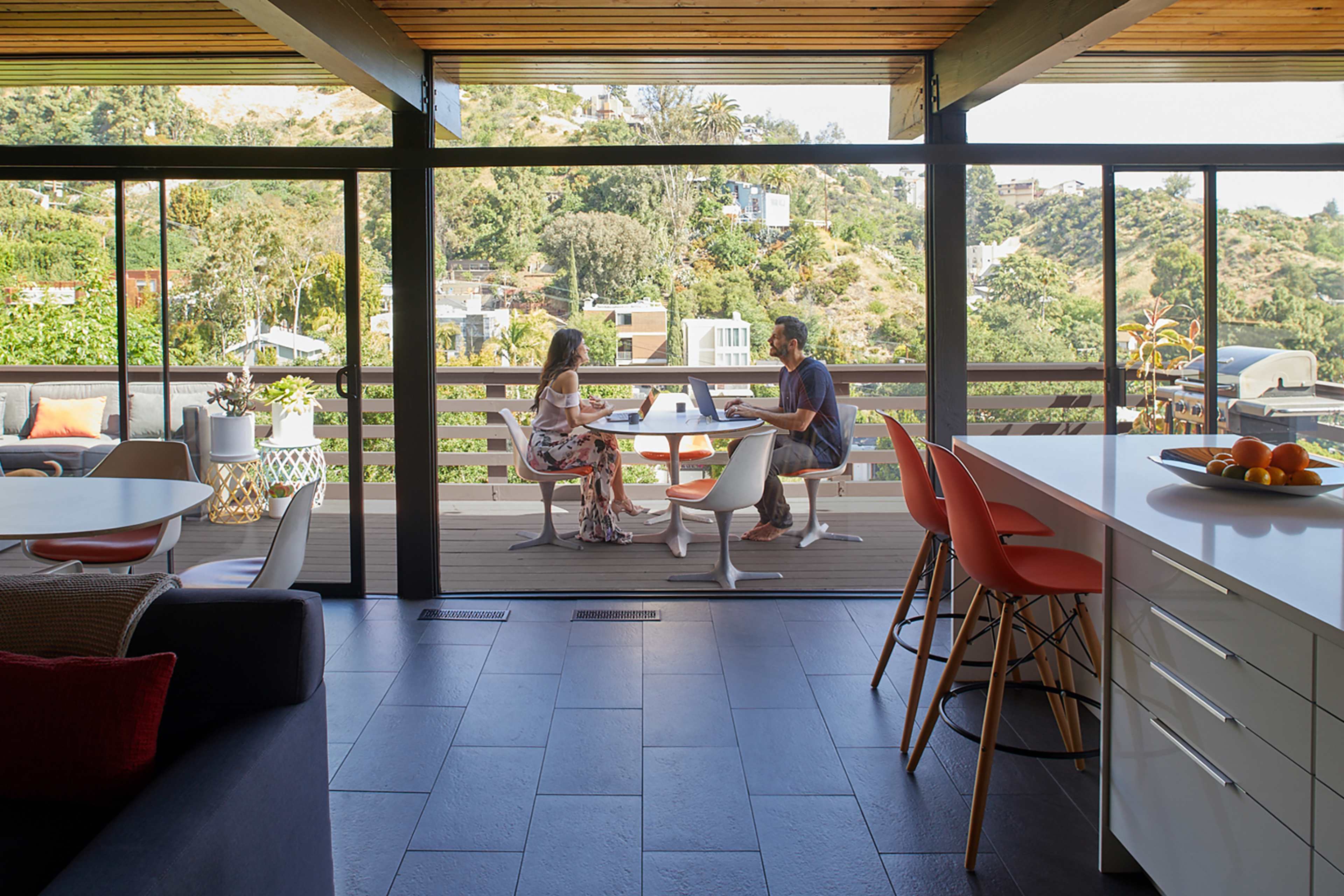 A couple sits at a table on a patio, working on a laptop with a scenic hillside view behind them.
