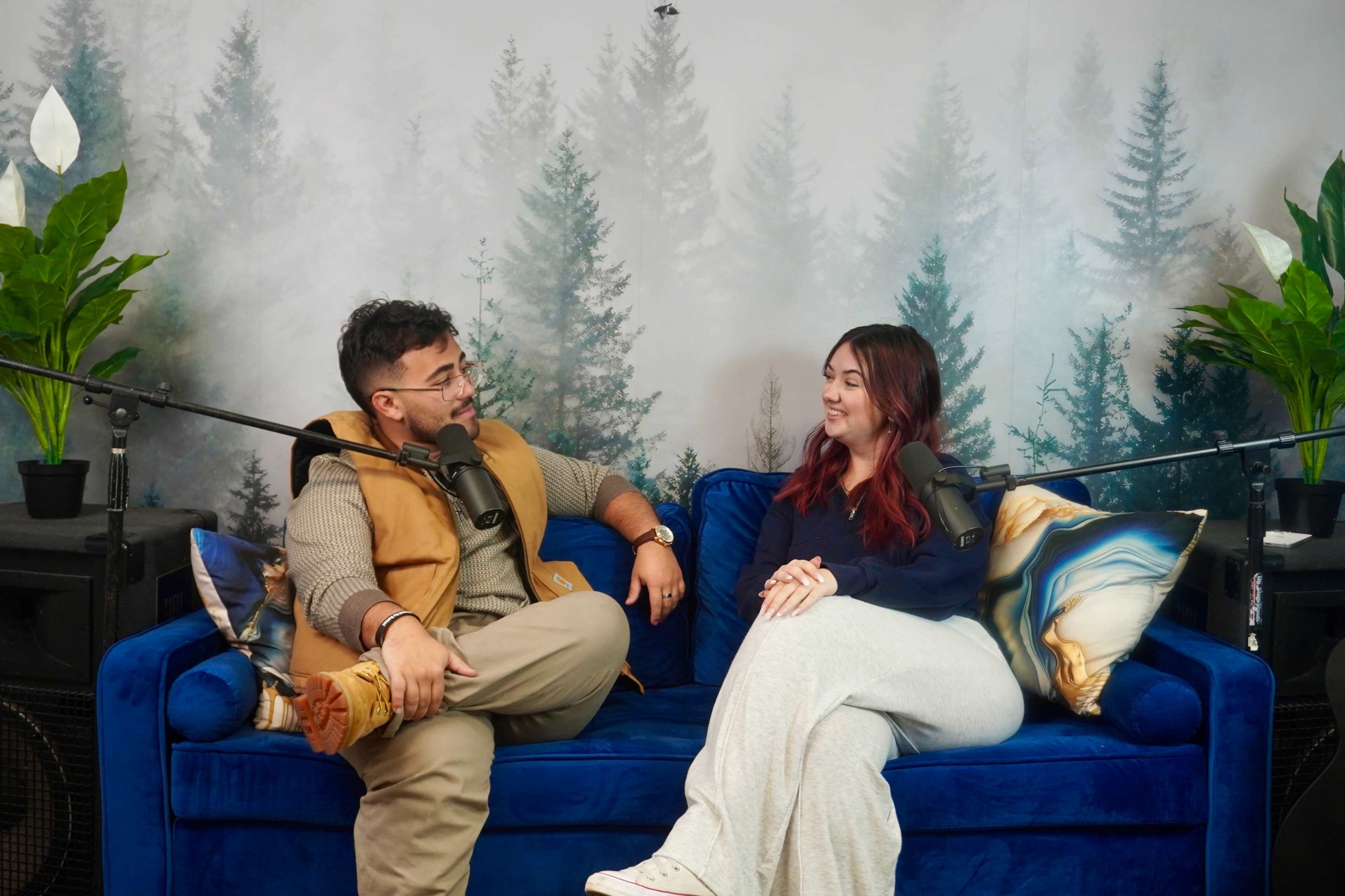 A man and a woman are seated on a blue couch in a studio adorned with plants and a forest-themed backdrop, engaged in conversation with microphones positioned in front of them.