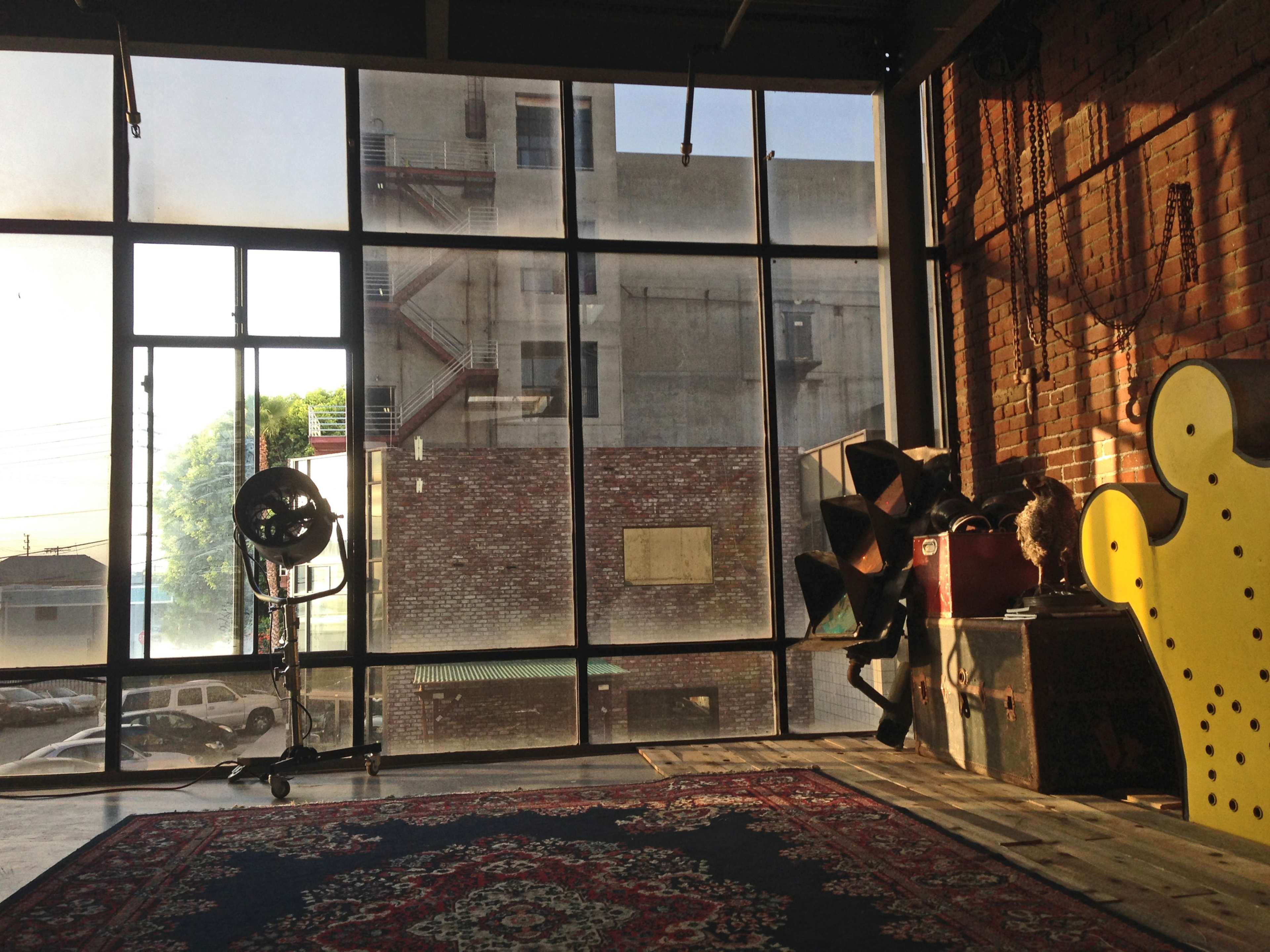 A spacious interior with large windows revealing a view of a building and parked cars outside, alongside a patterned rug and various objects on the floor.