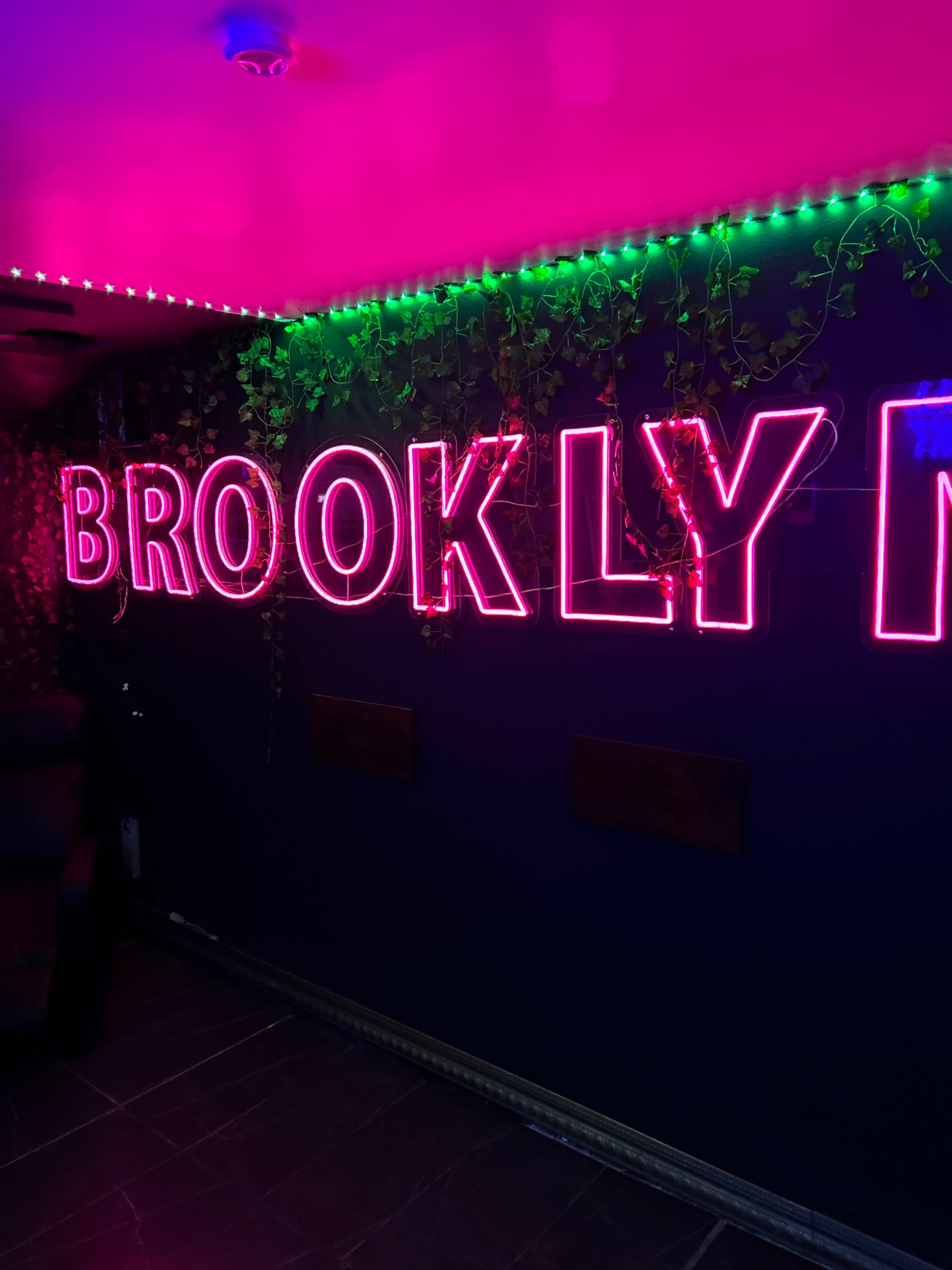 The image shows a brightly lit wall featuring the word "BROOKLYN" in large neon letters surrounded by green LED lights and decorative vines.