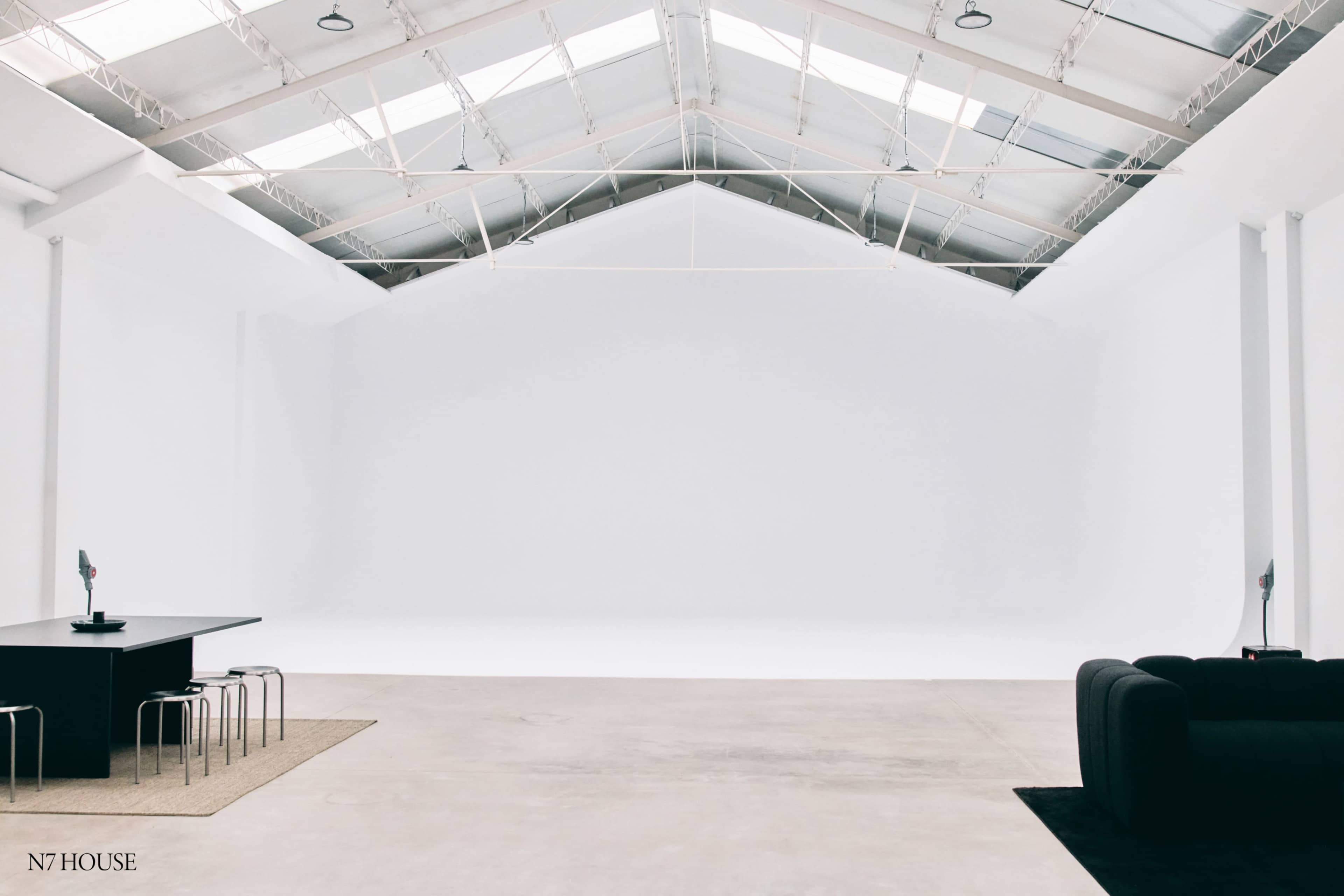 The image shows a spacious interior of a modern studio with a large, empty white wall and minimalist furniture, including a black table with chairs and a dark sofa.