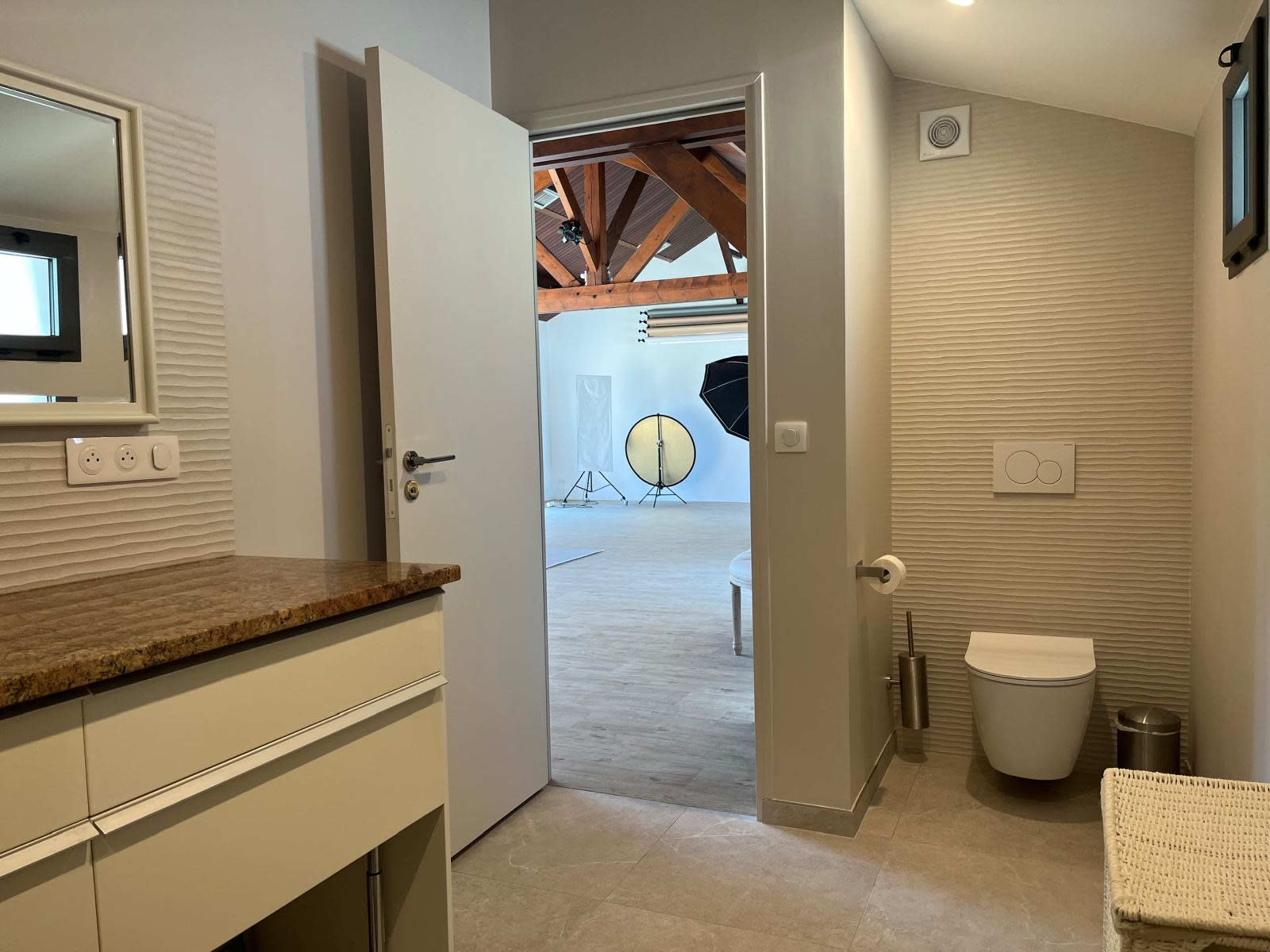 The image shows a bathroom with a modern design that connects to a spacious studio featuring wooden beams and photography equipment.