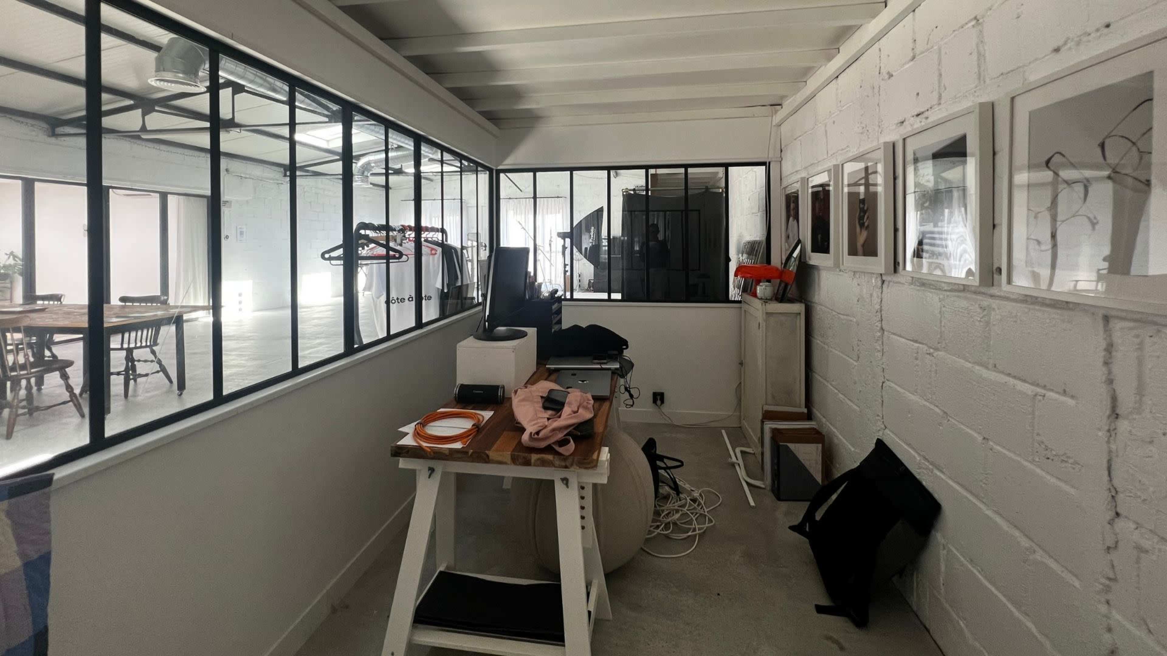 The image shows a modern workspace with a wooden table, a computer setup, and framed pictures on the walls, surrounded by large windows and a view of an open area.