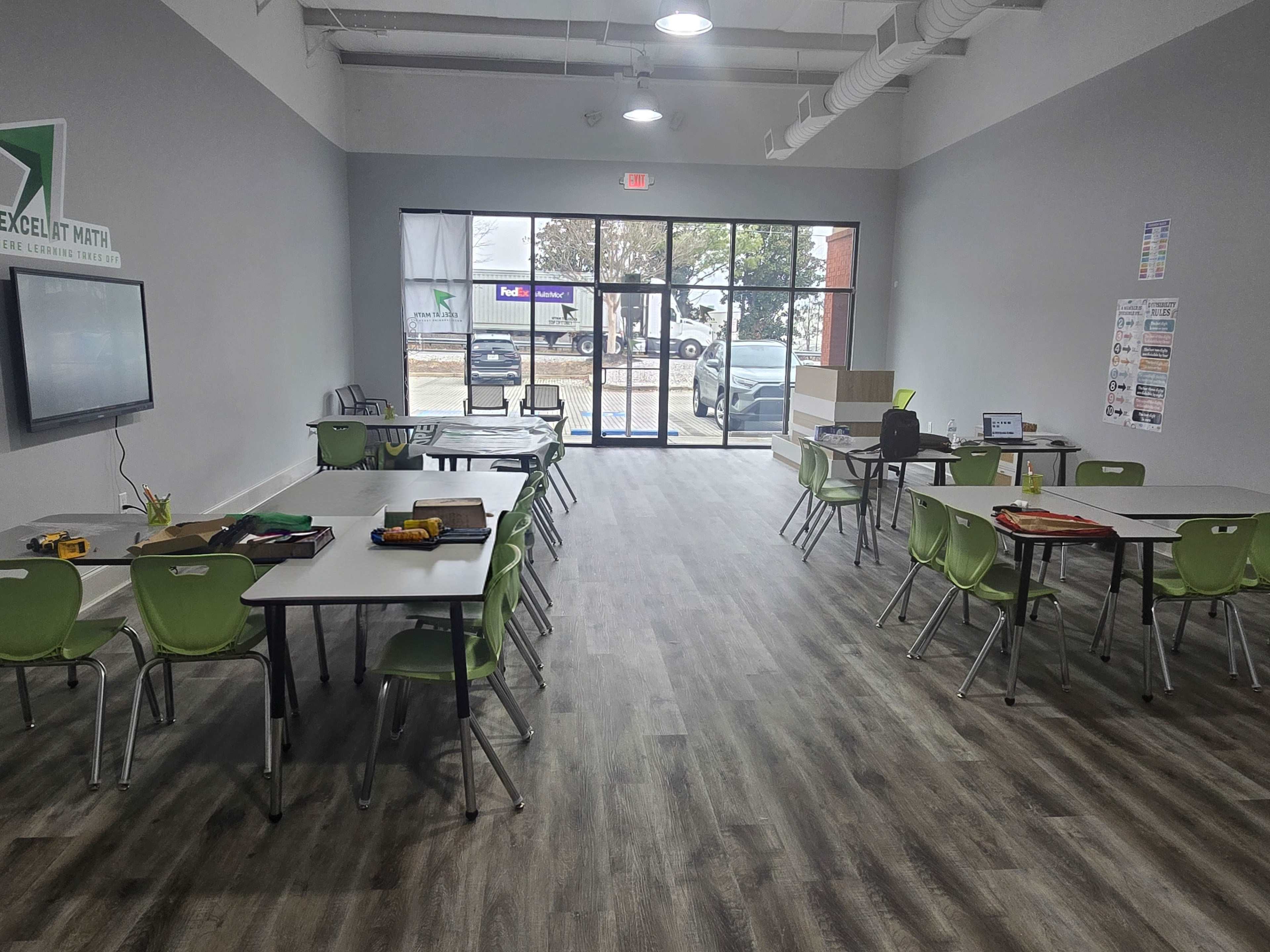 The image shows a bright, modern classroom with multiple tables and chairs arranged for an educational setting, featuring large windows that let in natural light.