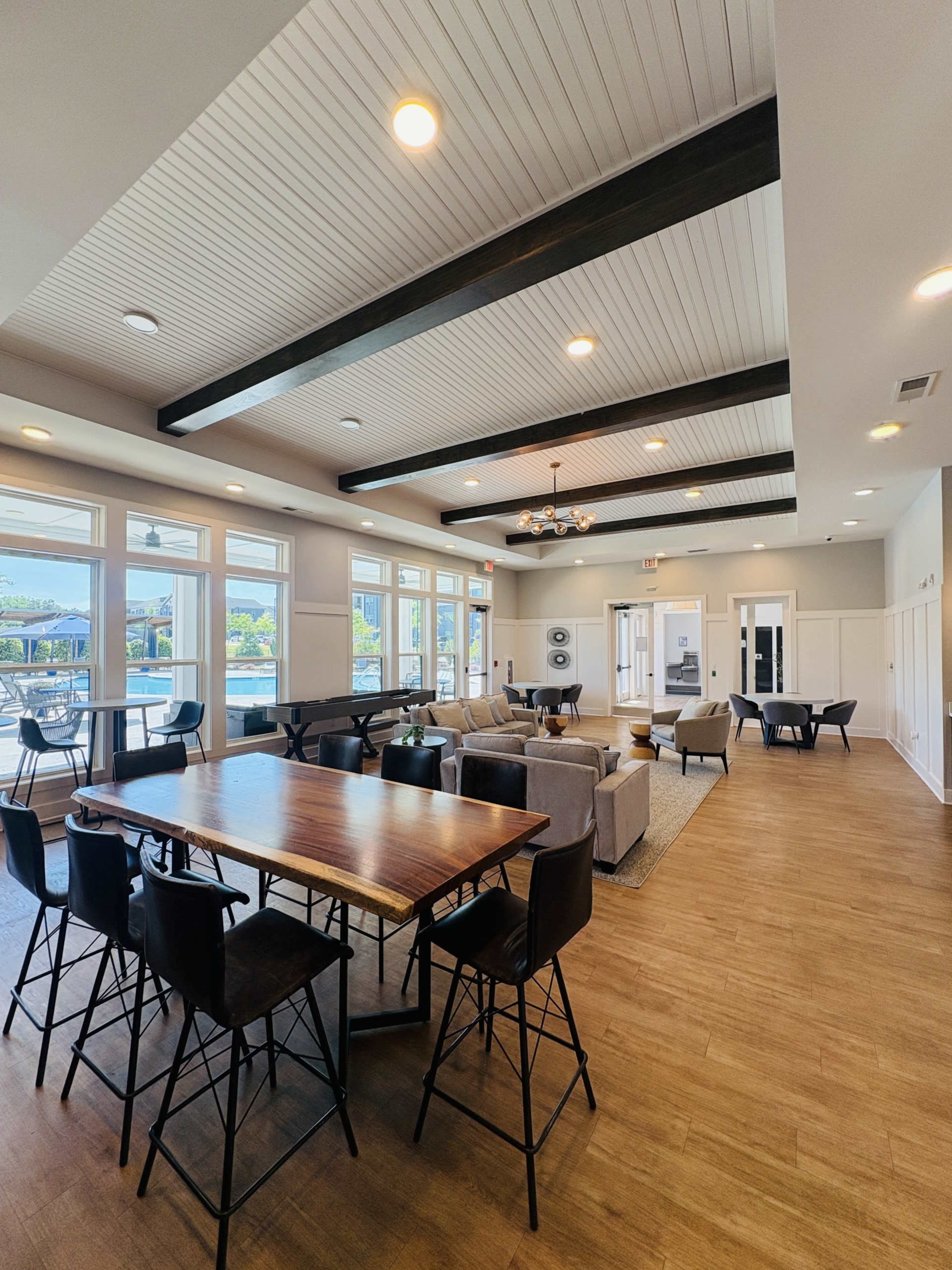 The image shows a modern, spacious lounge area with a large wooden table, several seating arrangements, and views of an outdoor space through large windows.