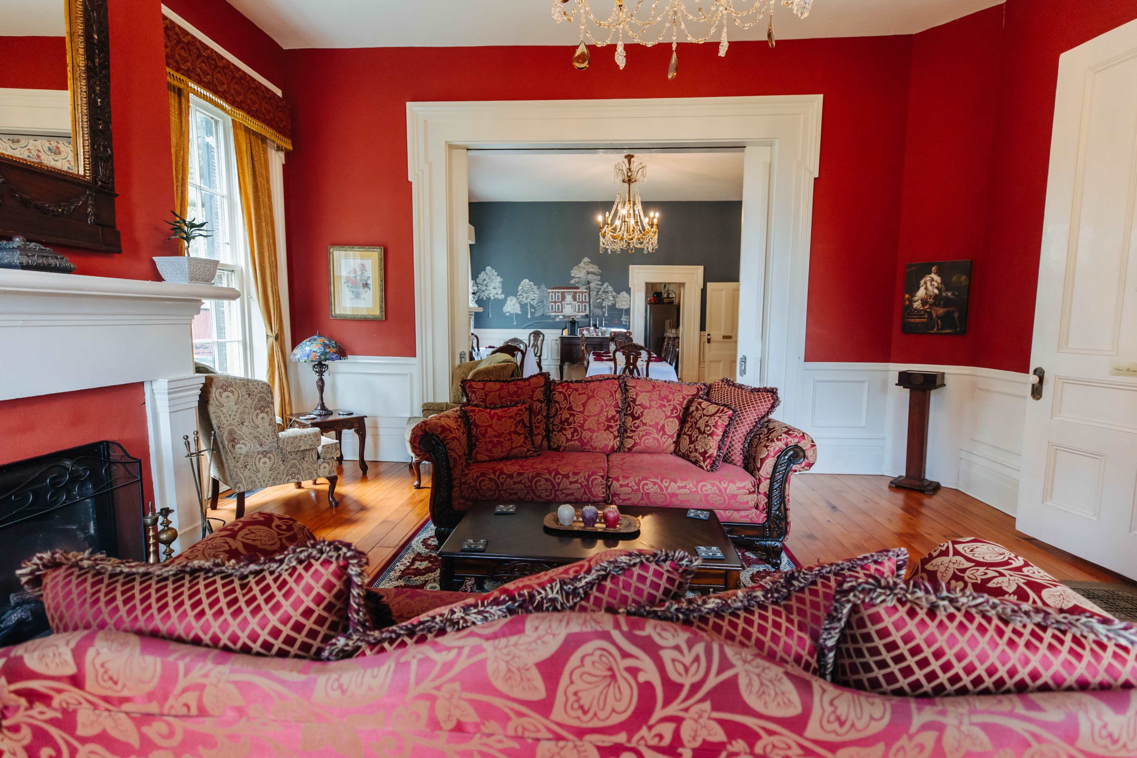 A richly decorated living room featuring a deep red wall, a patterned sofa, and a view into a dining area with a chandelier.