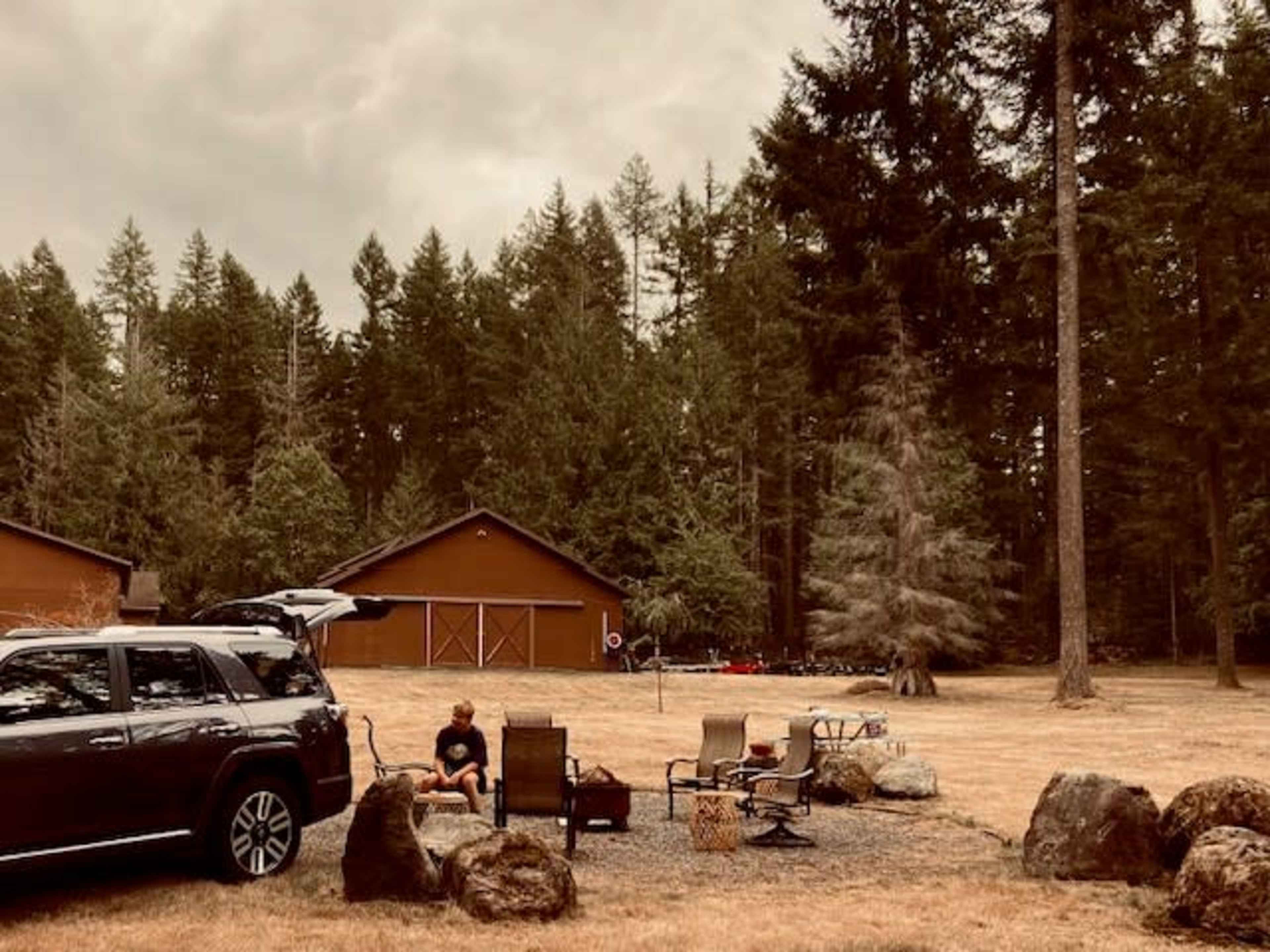 A person sits on a chair near a parked car in a grassy area surrounded by trees and rustic buildings.