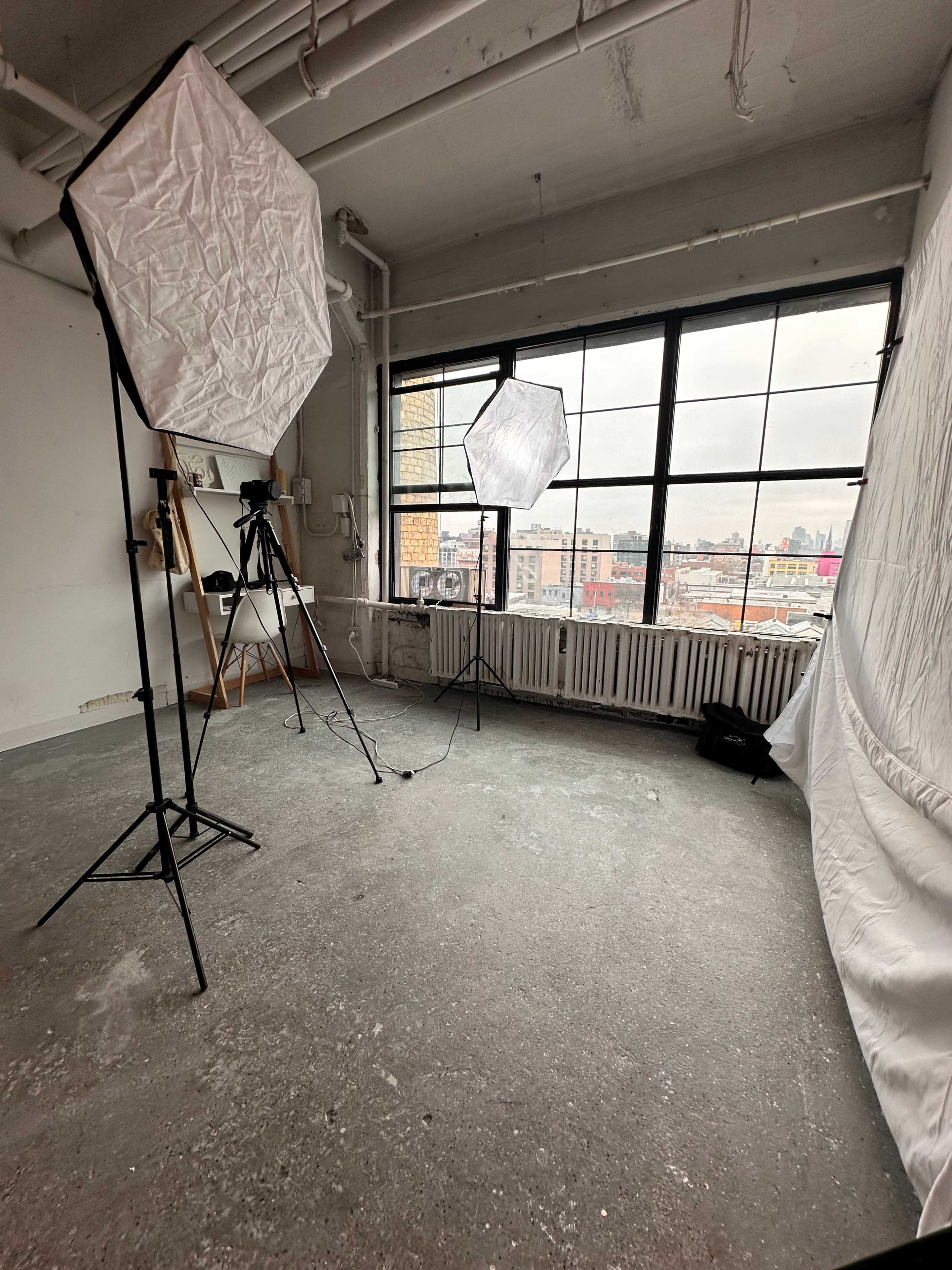 The image shows a photography studio with two softbox lighting setups, a camera on a tripod, and a white backdrop, all positioned in front of large windows overlooking a cityscape.