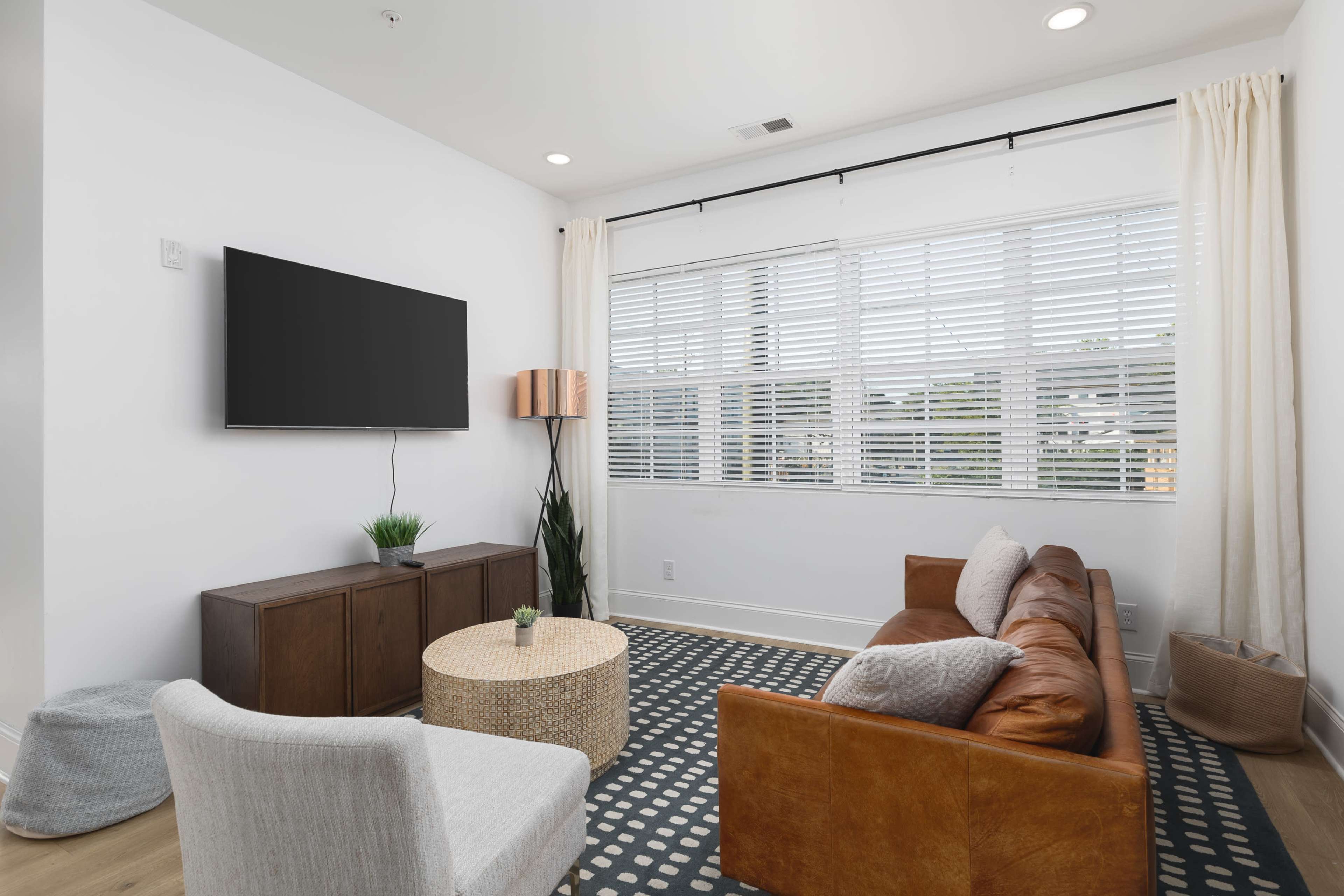 A living room features a television mounted on the wall, a brown leather sofa, a round coffee table, and a window with white blinds.
