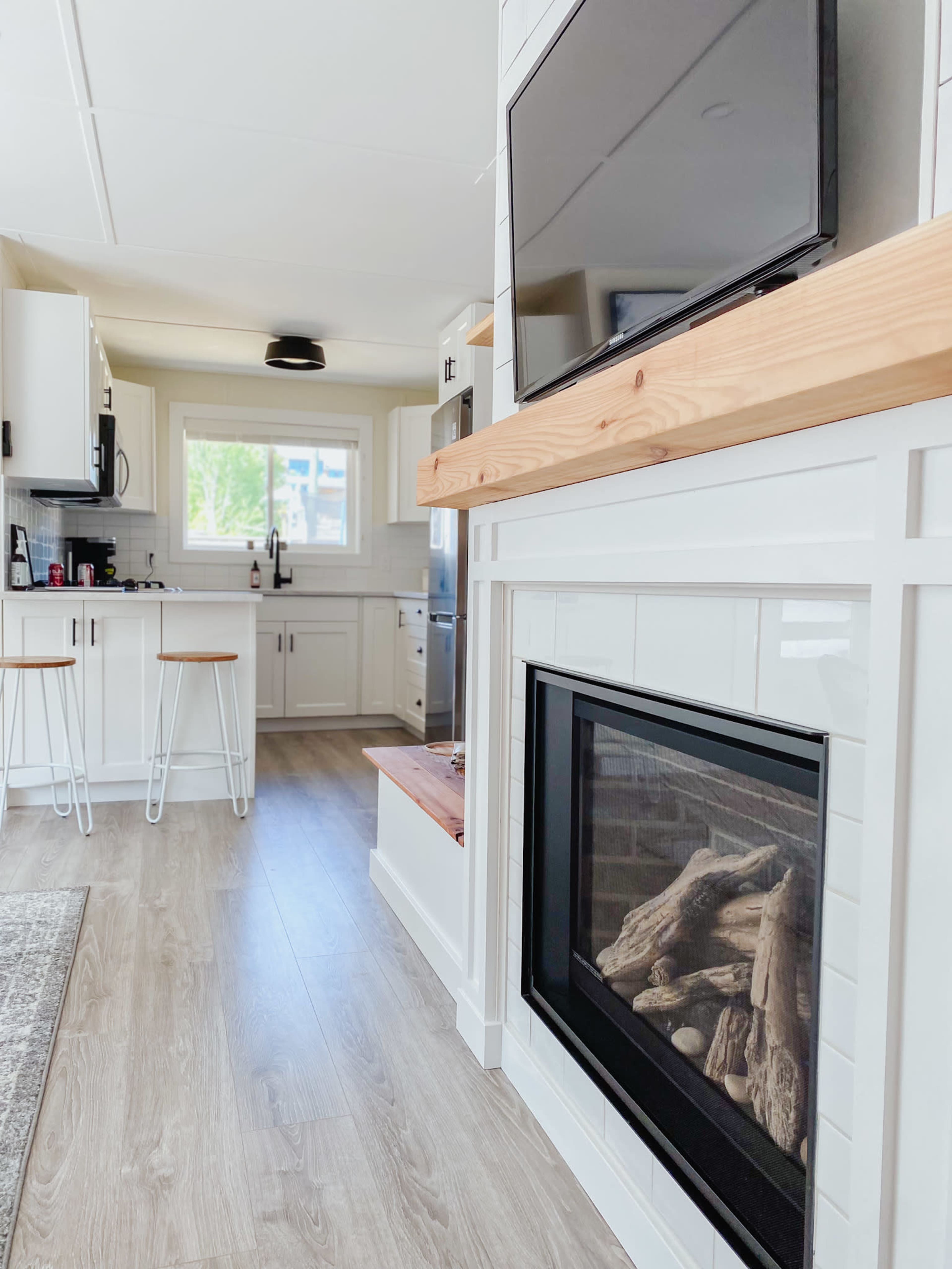 A modern living space features a sleek fireplace below a mounted television, with a kitchen area and minimalist bar stools in the background.